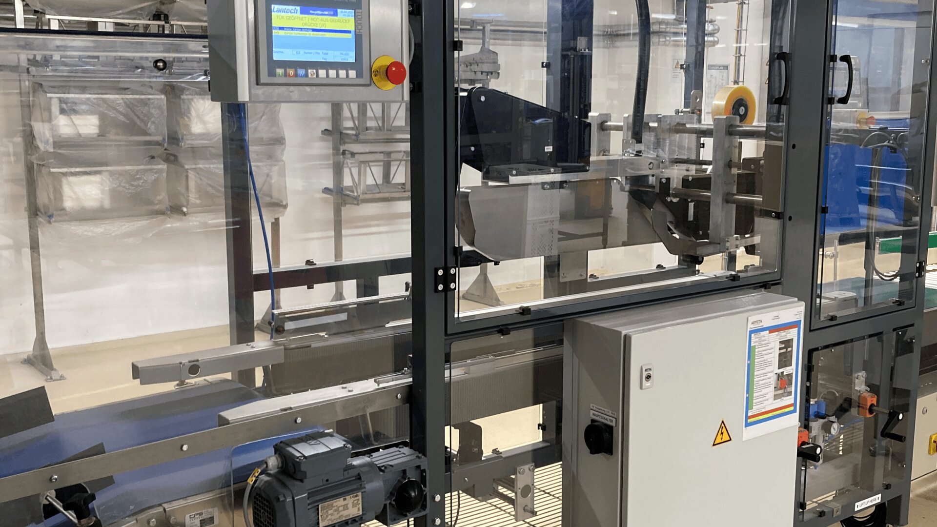 Eine Industrieverpackungsmaschine von Lantech steht in einer sauberen, gut beleuchteten Halle. Die Maschine verfügt über ein Bedienfeld mit Touchscreen-Display, Förderbänder und transparente Sicherheitspaneele.
