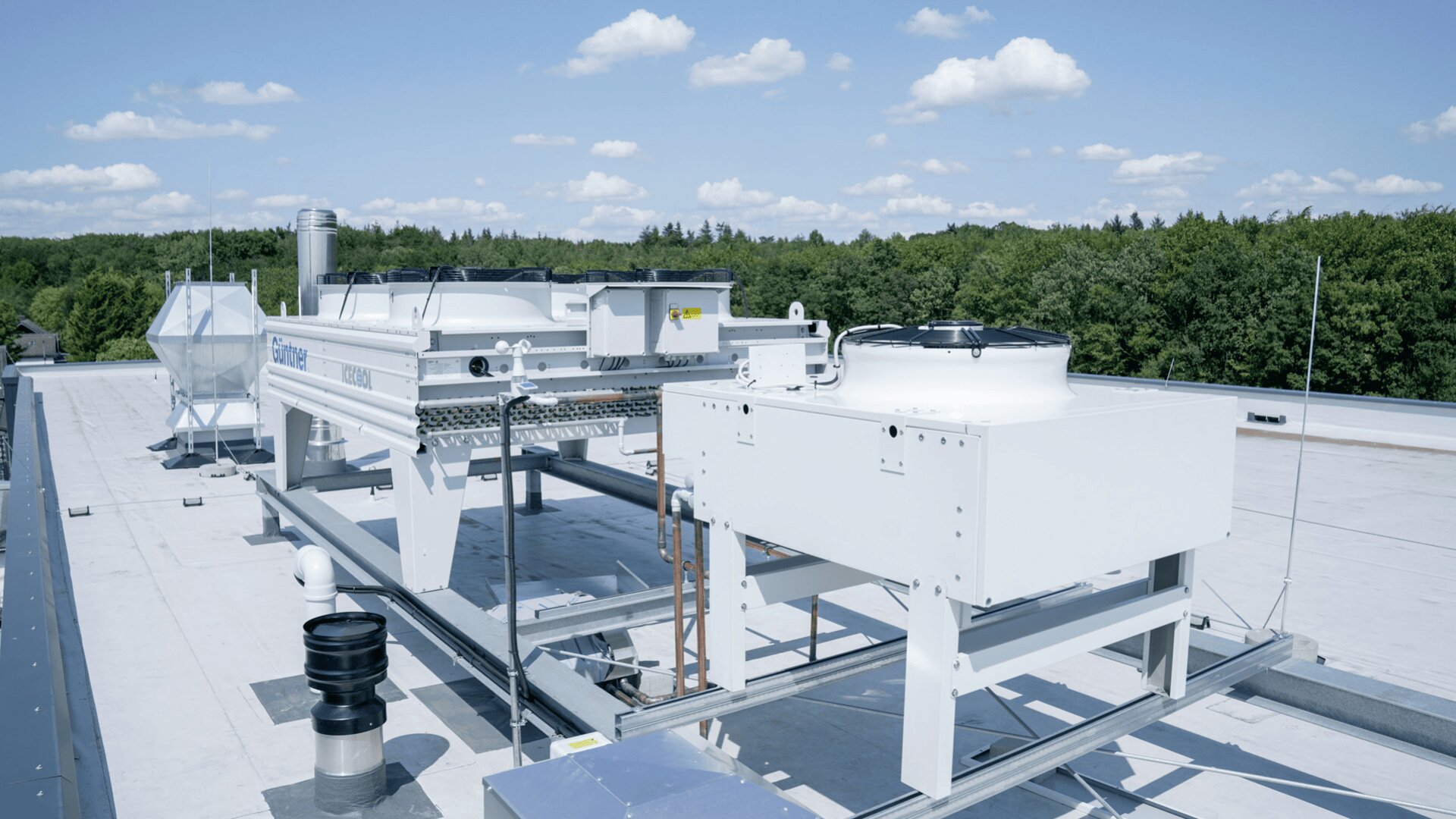 Große industrielle HVAC-Einheiten sind auf einem flachen Dach unter einem klaren blauen Himmel mit vereinzelten Wolken installiert, umgeben von Bäumen im Hintergrund.