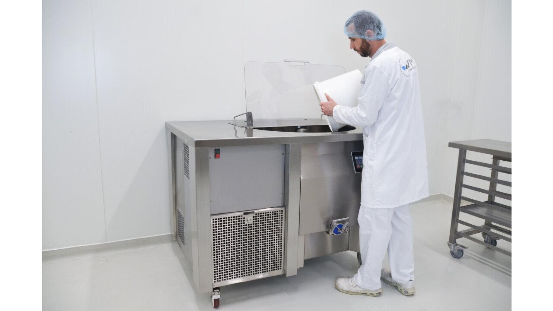 Eine Person in weißer Schutzkleidung und einem Haarnetz schüttet einen weißen Behälter in einen großen Industriemischer aus Edelstahl in einer sauberen, weißwandigen Anlage.