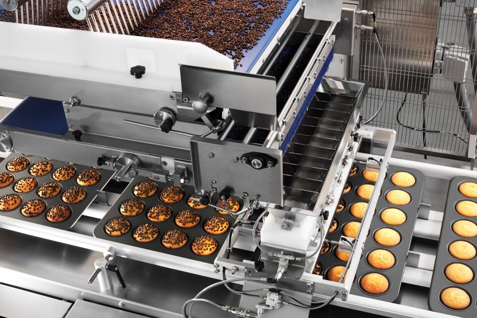 Industrial baking machine dispensing chocolate chips onto trays of muffins on a conveyor belt in a factory setting. Some muffins are plain, while others are being topped with chocolate chips.