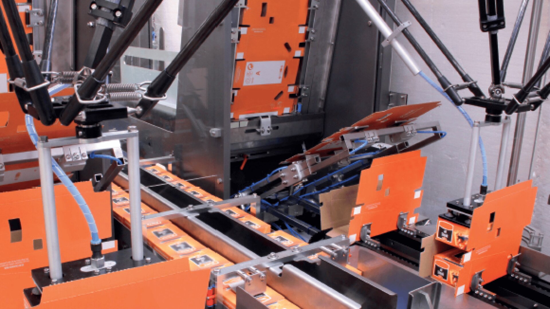 Several robotic arms are assembling and packing orange cardboard boxes on an automated production line in a factory setting.