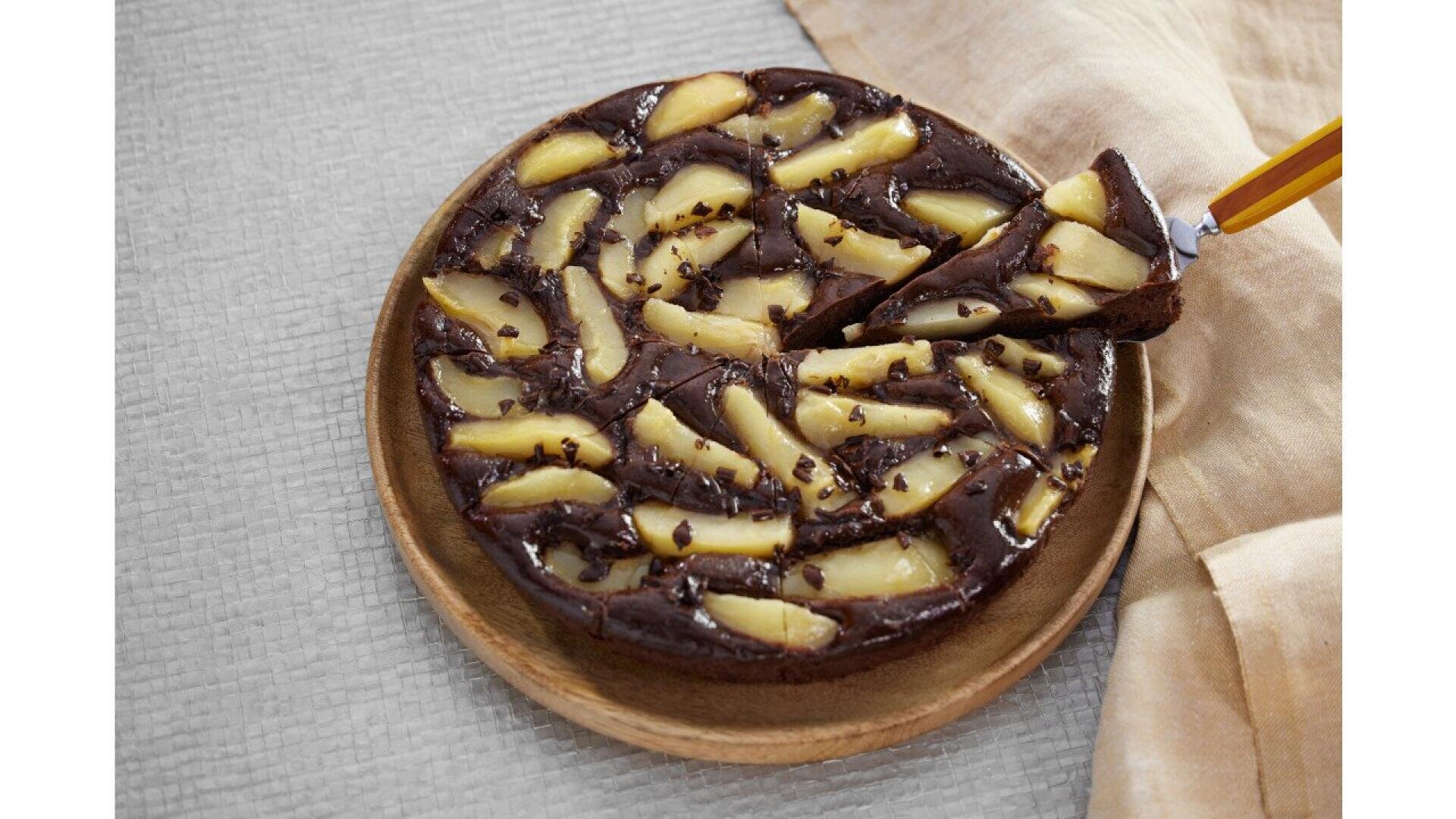 A round chocolate cake topped with slices of pear and sprinkled with chocolate chips, placed on a wooden plate. A spatula is lifting one slice. A beige cloth lies beside the cake on a textured surface.