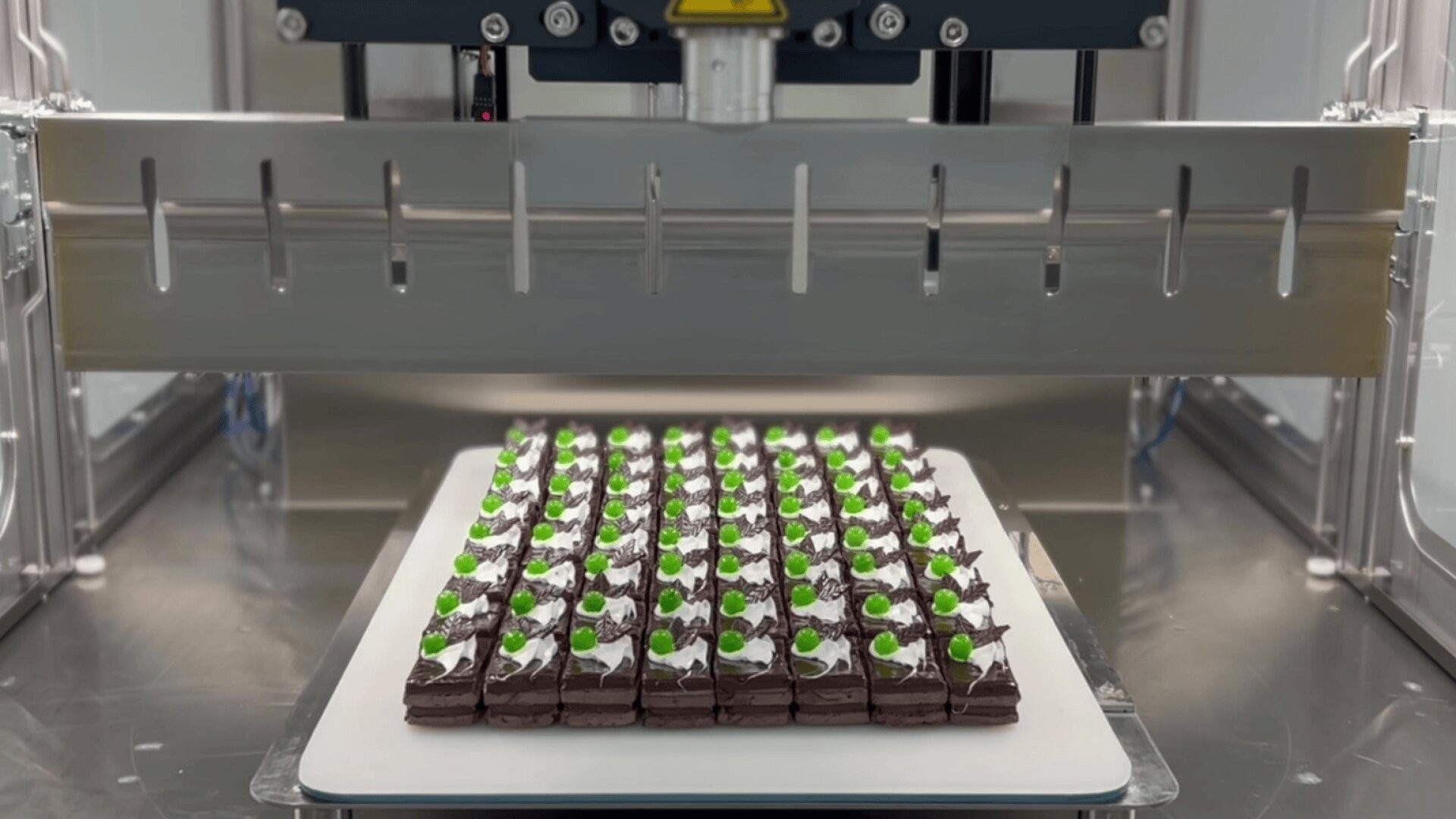 Ein Tablett mit fein säuberlich angeordneten Schokoladenquadraten, die mit weißer Sahne und grünen Verzierungen überzogen sind, liegt unter einer industriellen Schneidemaschine, bereit, in einzelne Stücke geschnitten zu werden.