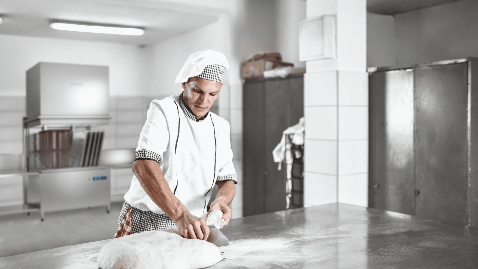 Ein Bäcker in weißer Uniform und mit Hut knetet Teig auf einer bemehlten Metallarbeitsfläche in einer sauberen, modernen Bäckereiküche mit Edelstahlgeräten und gefliesten Wänden im Hintergrund.