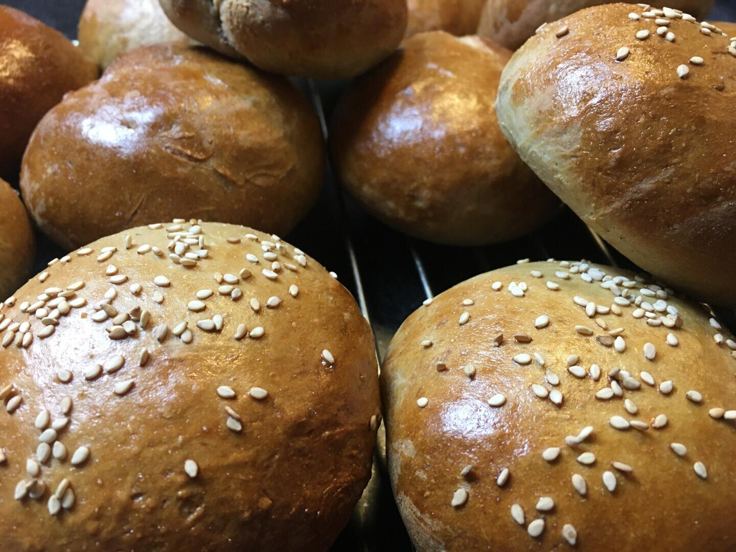 Close-up of several golden-brown hamburger buns, some topped with sesame seeds, arranged closely together in a pile.