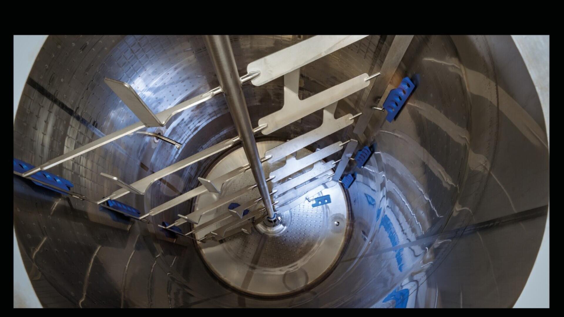 Interior view of a large, shiny stainless steel industrial mixing tank with metal paddles and blue brackets, designed for blending or processing materials.