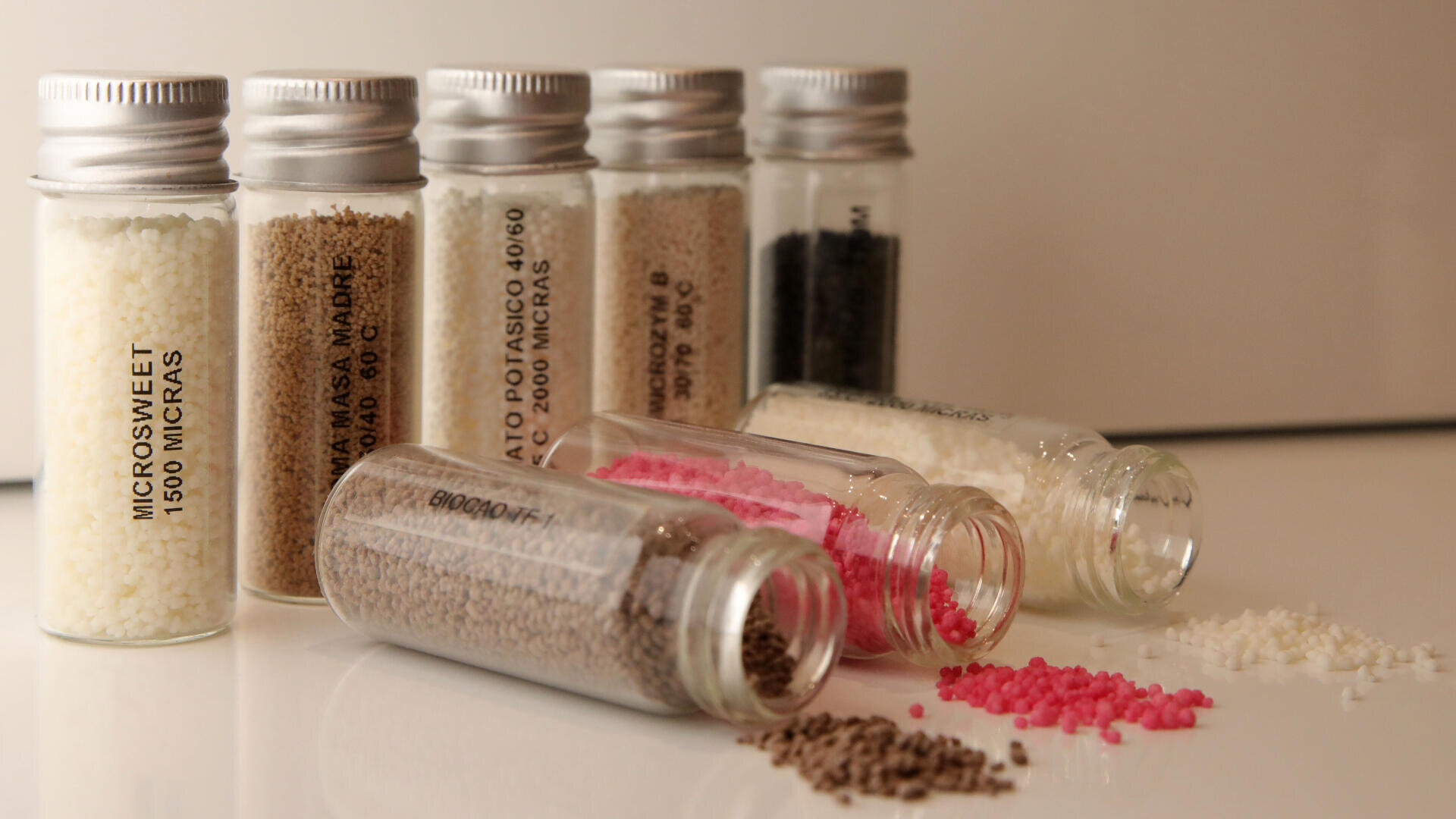 Six small glass vials, some upright and some tipped over, contain different colored granular substances, including white, brown, pink, and black, with some granules spilled onto a white surface.