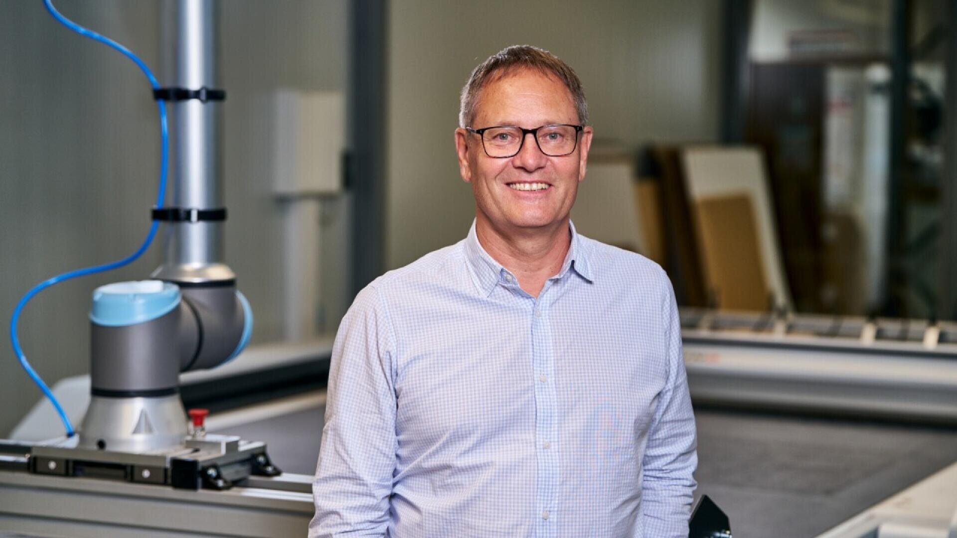 A middle-aged man wearing glasses and a light blue button-up shirt smiles while standing in an industrial or manufacturing setting with equipment and machinery in the background.