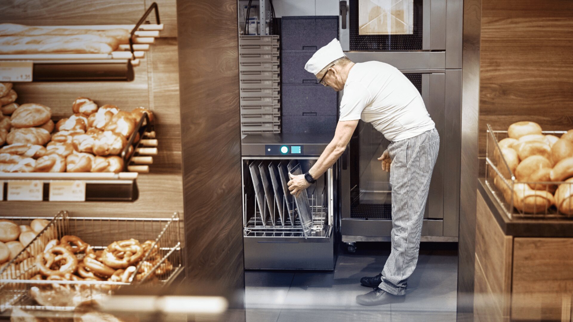Ein Bäcker mit weißer Mütze und weißem Hemd lädt Tabletts in eine industrielle Spülmaschine in einer Bäckerei, umgeben von Regalen mit verschiedenen Brotsorten und Gebäck.