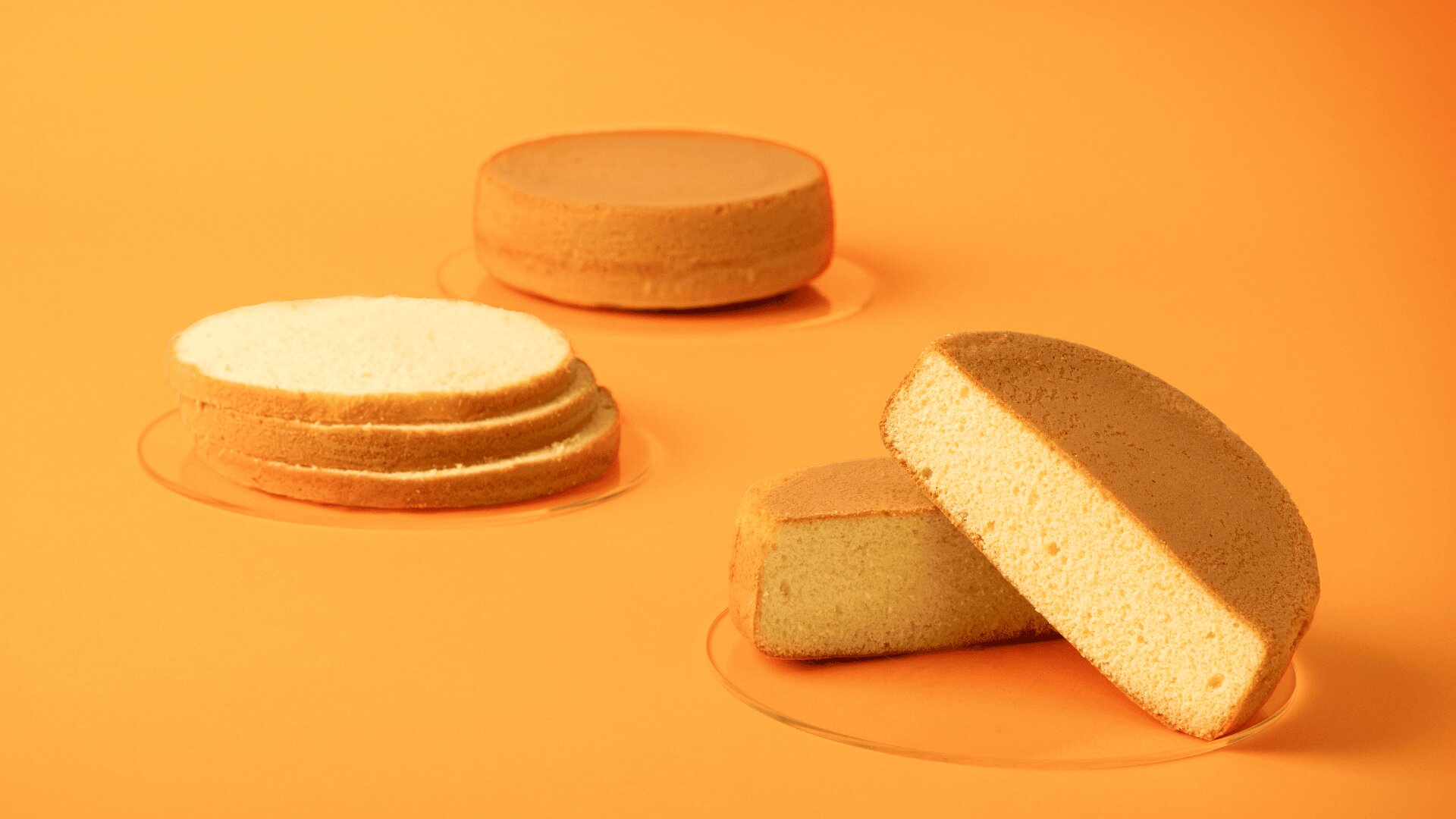 Three plain sponge cakes on an orange surface: one whole, one sliced into three layers, and one cut in half with the cut side facing forward.