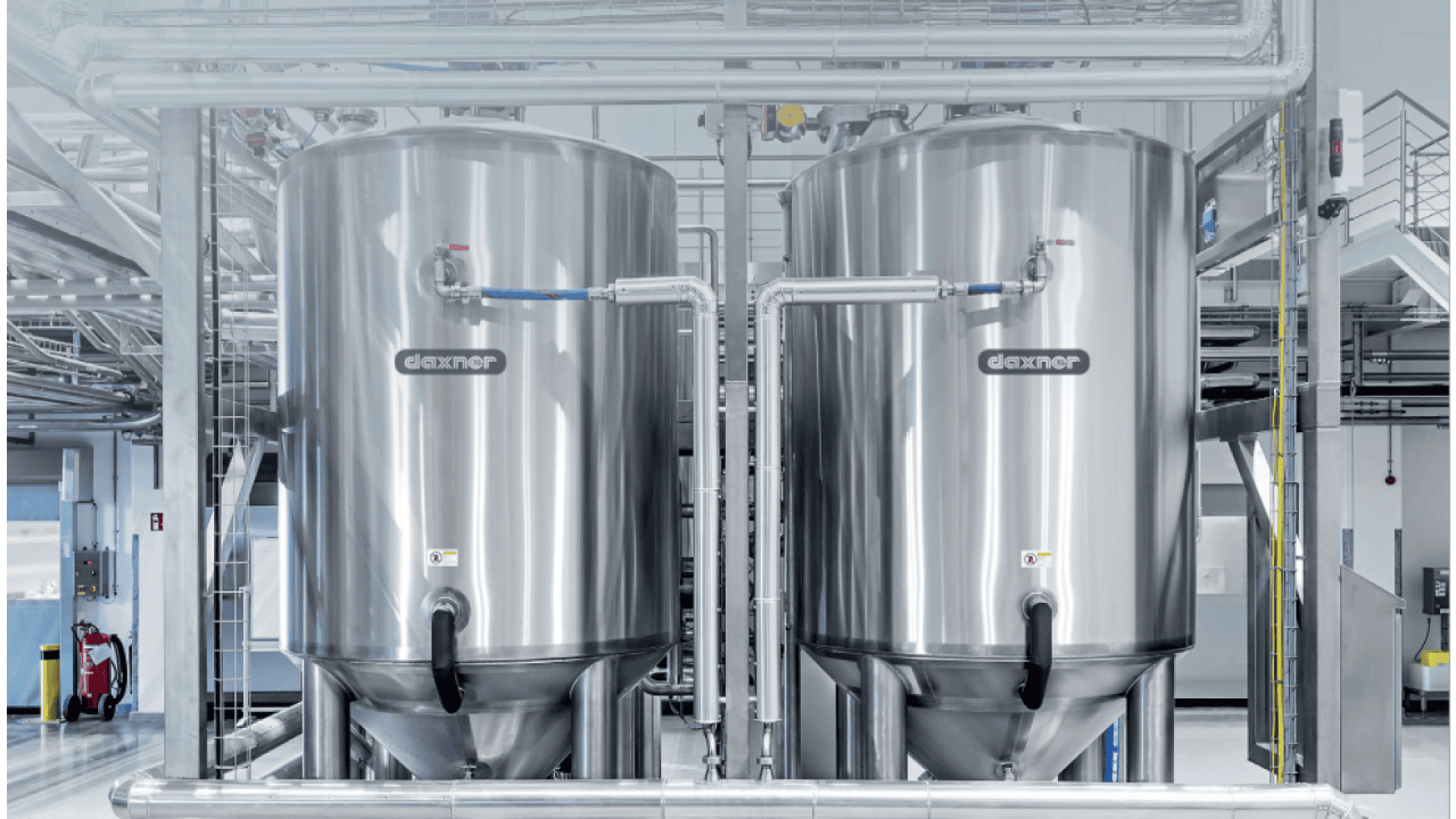 Two large stainless steel tanks connected by pipes stand in a clean, industrial facility with metal railings and ladders in the background. The tanks are marked with the brand name GASTNER.