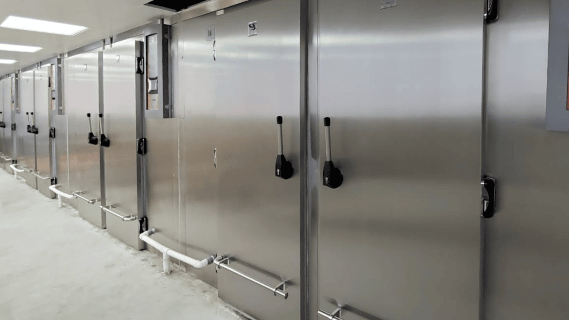 A row of large, stainless steel industrial refrigerators or cold storage units with black handles and visible pipes along the bottom, lined up in a clean, brightly lit facility.