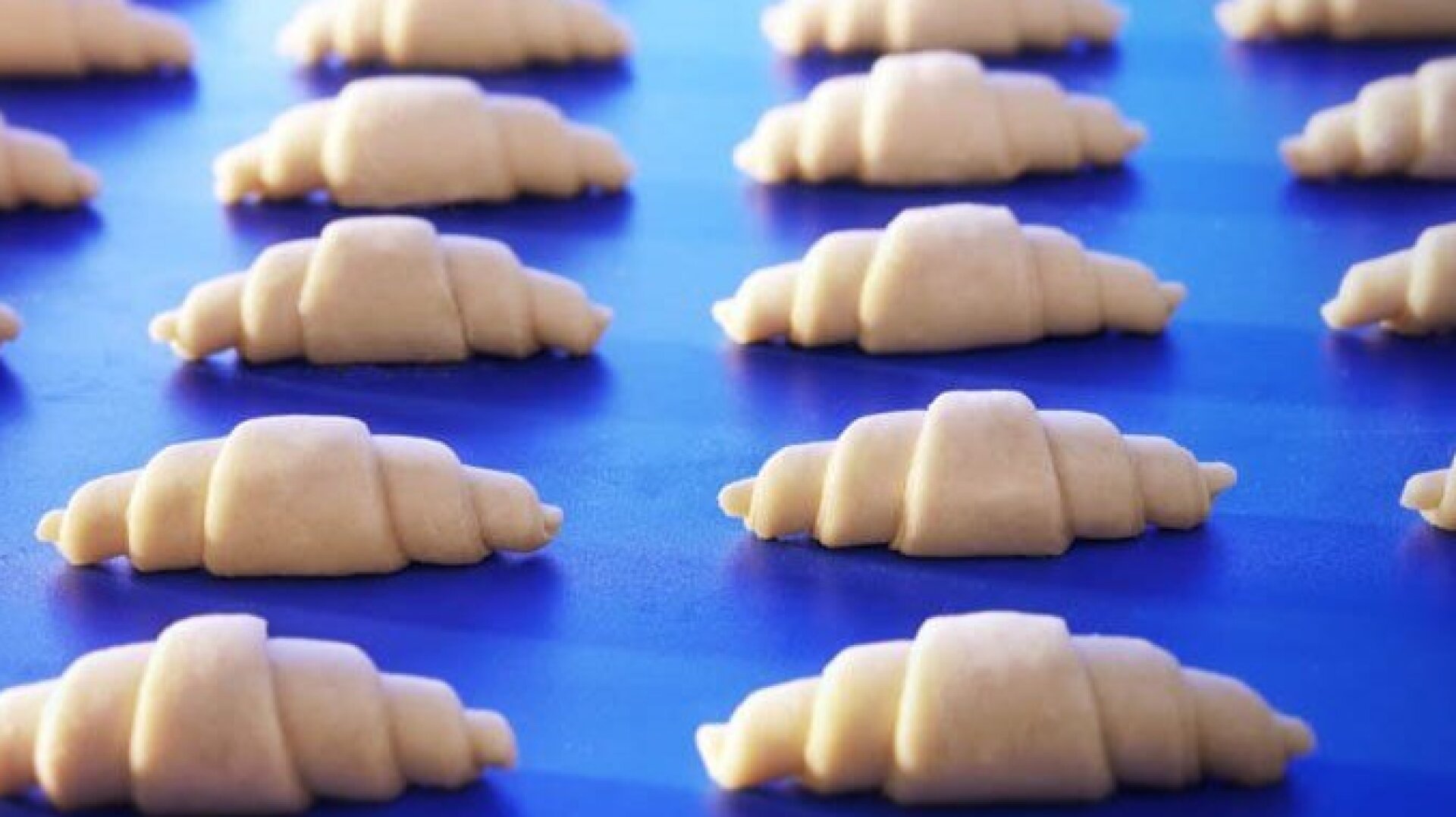 Rows of unbaked croissant dough pieces are neatly arranged on a blue surface, ready to bake.