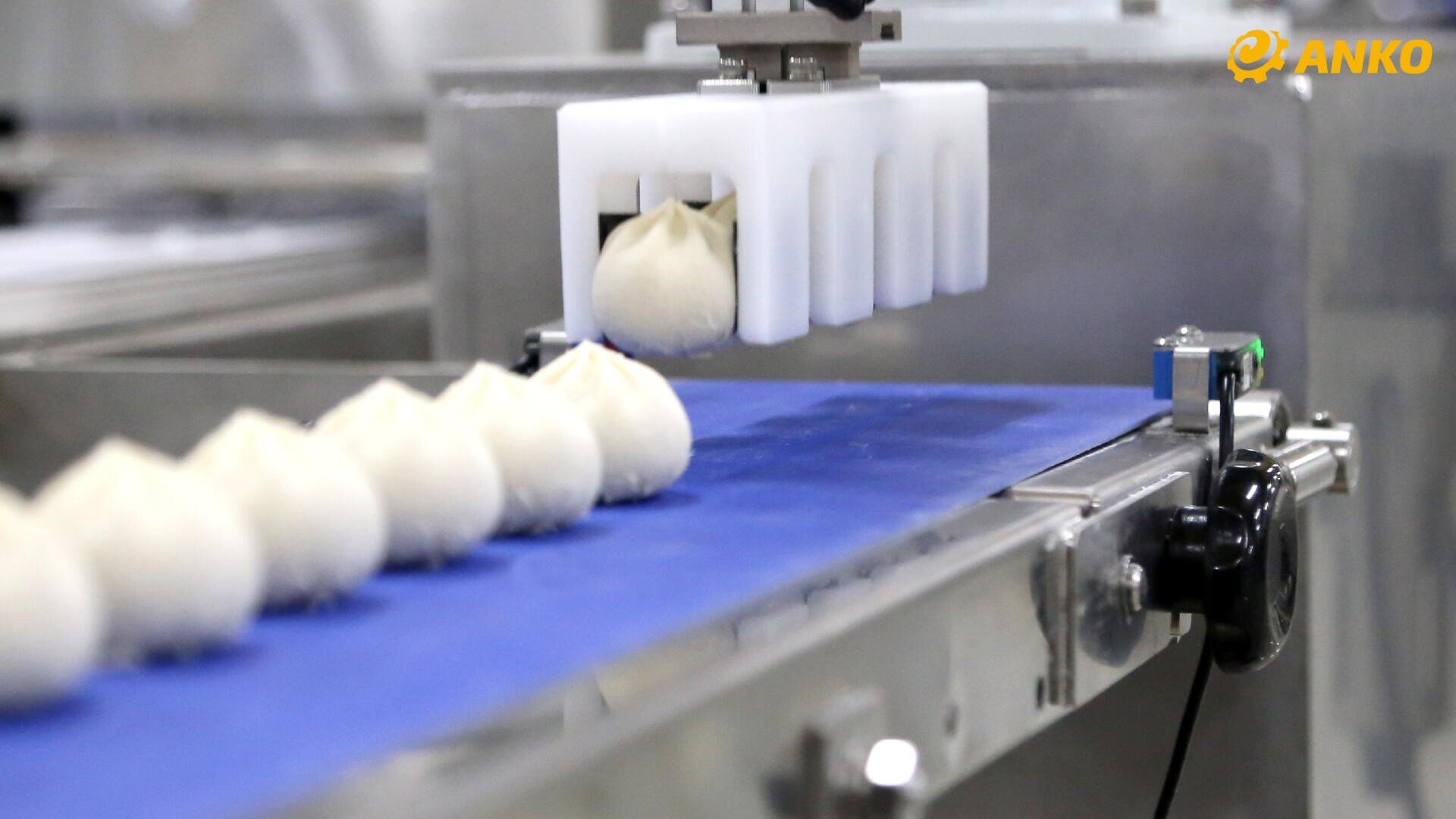 Nahaufnahme einer Lebensmittelverarbeitungsmaschine, die gedämpfte Brötchen auf ein blaues Förderband in einer Fabrik legt, mit mehreren aufgereihten Brötchen und dem ANKO-Firmenlogo in der oberen rechten Ecke.
