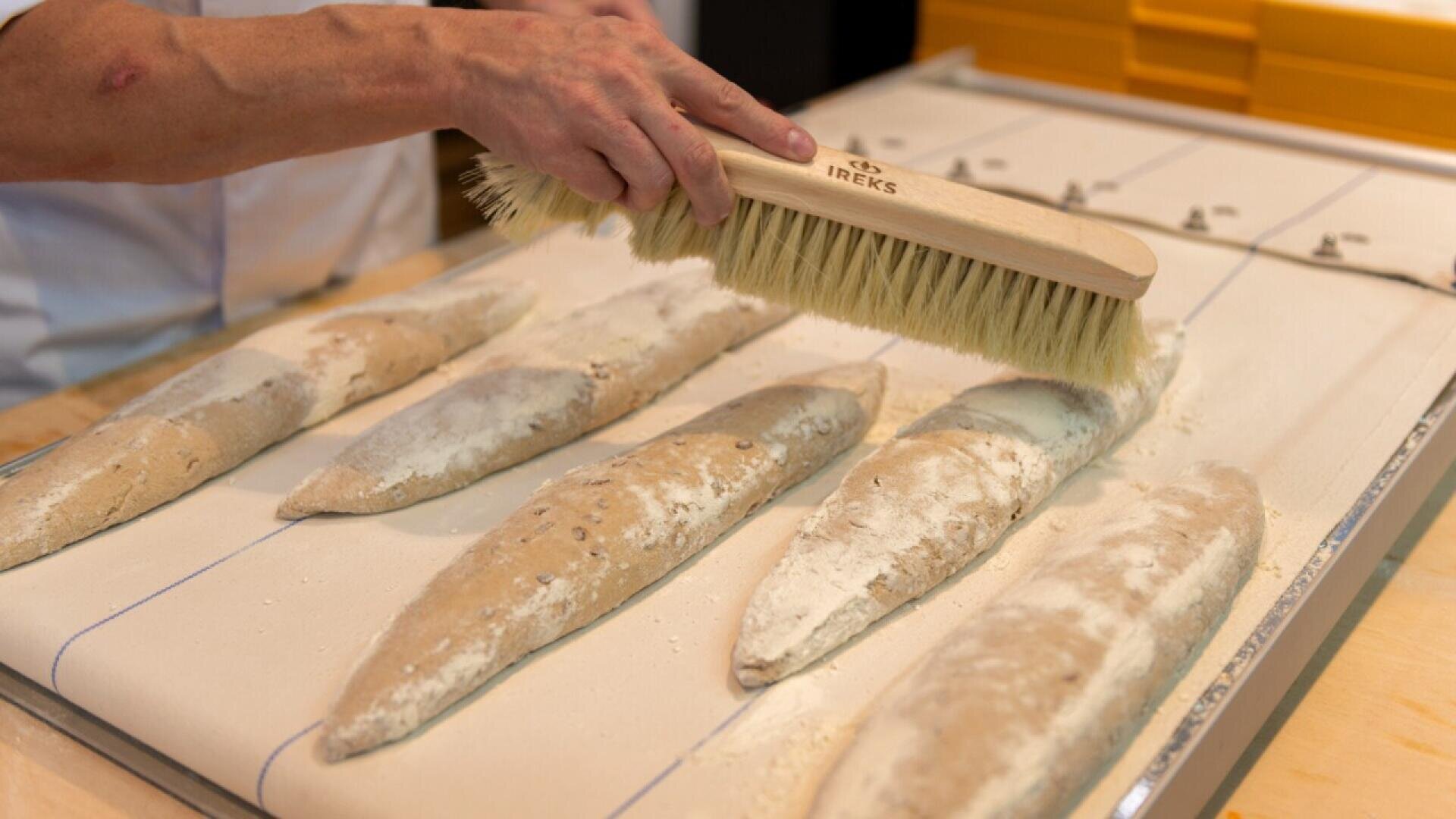 A person uses a large brush to dust flour off several uncooked baguettes lined up on a cloth-covered surface in a bakery setting.