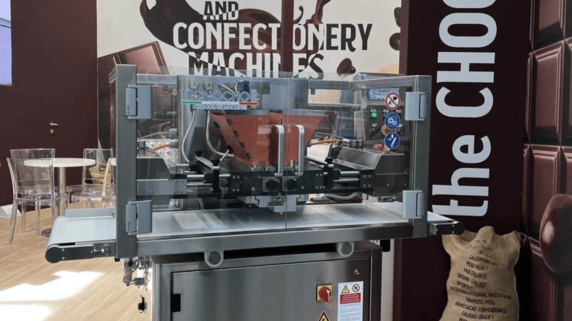 A chocolate processing machine with transparent protective shielding, displayed in front of a backdrop that reads Confectionery Machines and the CHOC, with images of chocolate bars and a burlap sack.