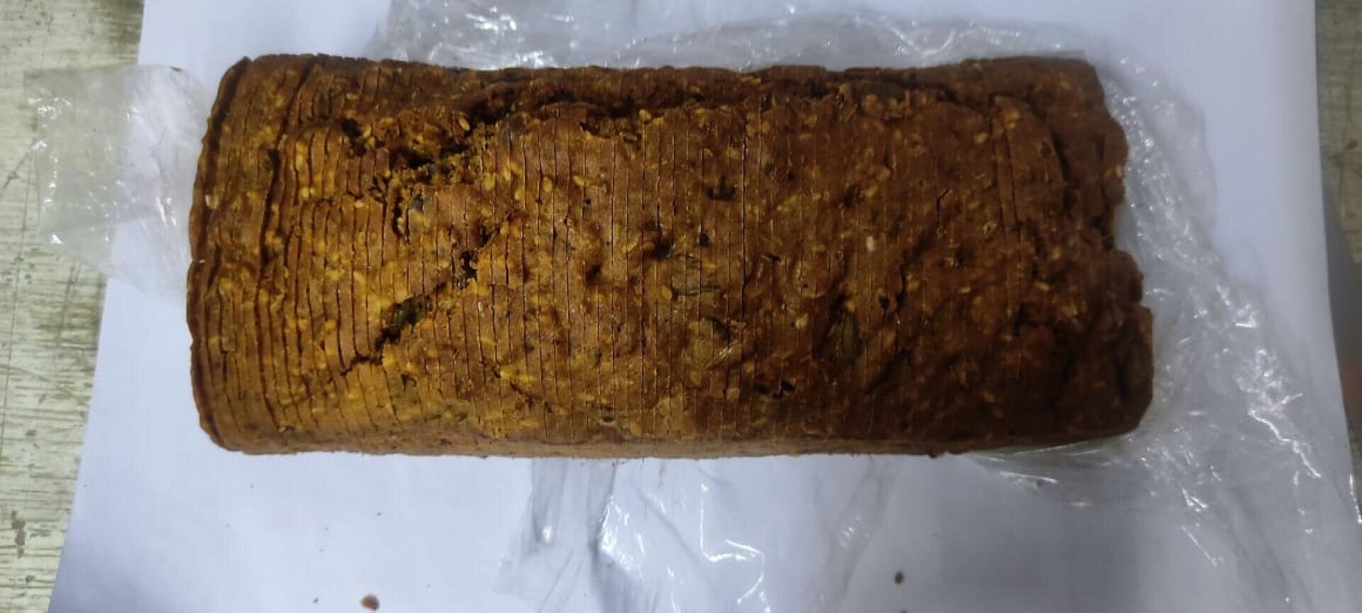 A loaf of brown, crusty bread with a rough texture rests on a piece of clear plastic wrap over a white sheet of paper.