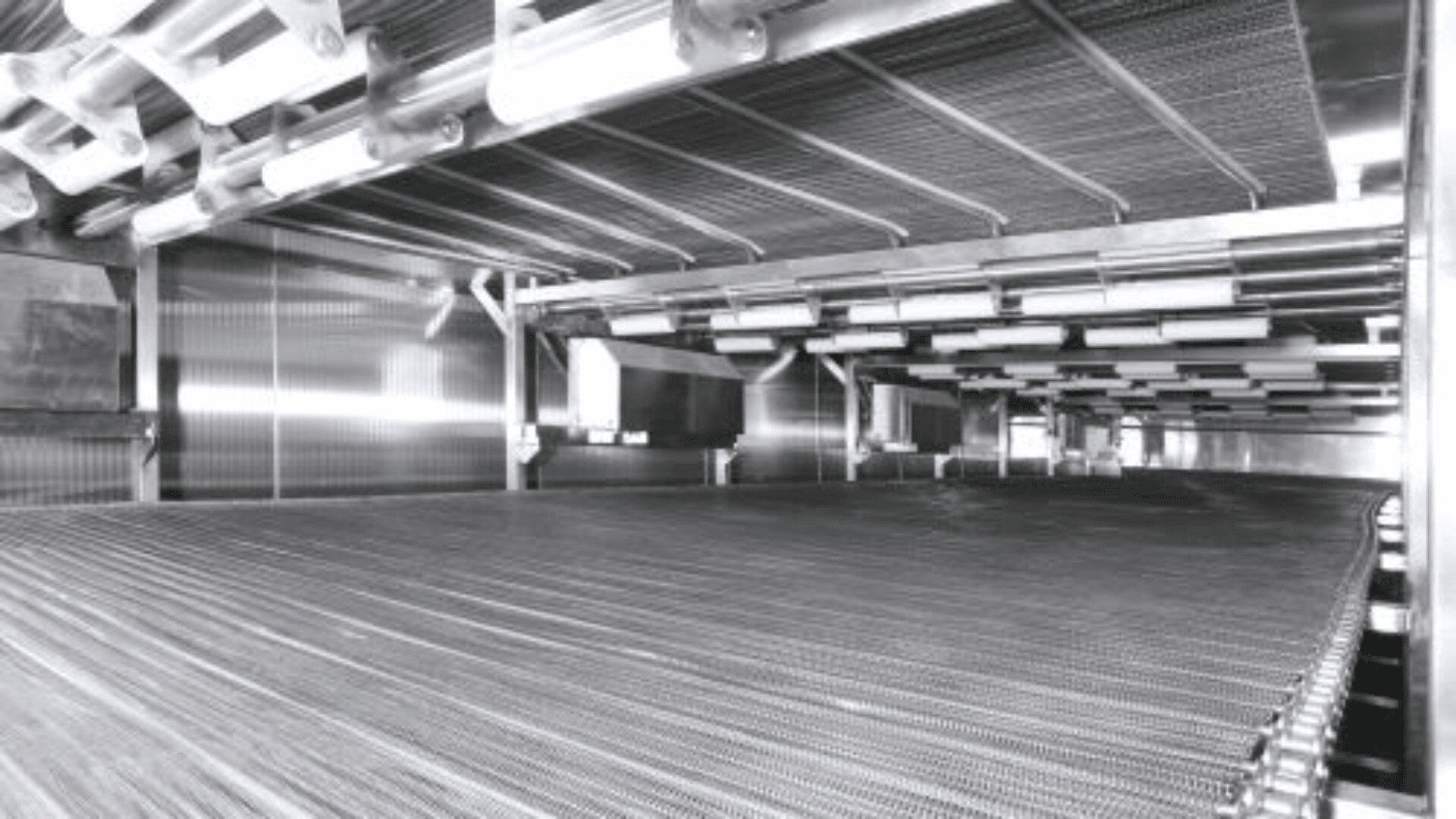 A large industrial conveyor system with metal rollers and a mesh surface inside a spacious, metallic factory setting, viewed from a low angle.
