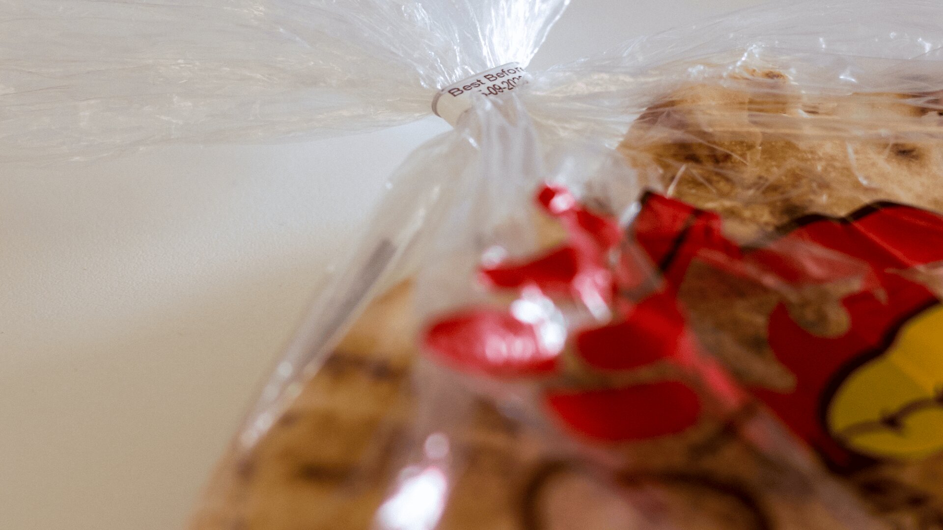 A close-up of a plastic bread bag sealed with a twist tie. The twist tie displays a Best Before date. Part of the bread and some red and yellow print on the bag are visible in the foreground.