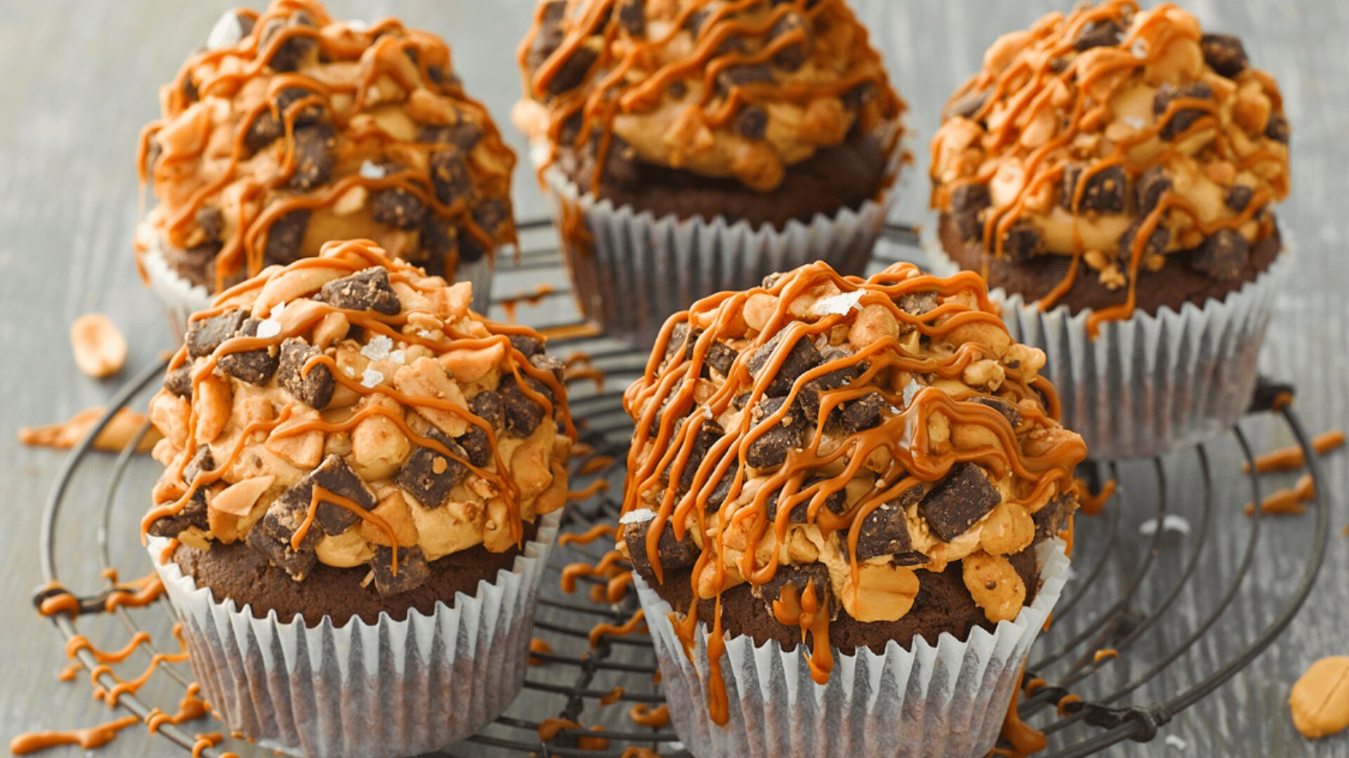 Five chocolate cupcakes topped with chopped nuts, chocolate chunks, and drizzled with caramel sit on a wire cooling rack. The background is a neutral, slightly blurred surface.