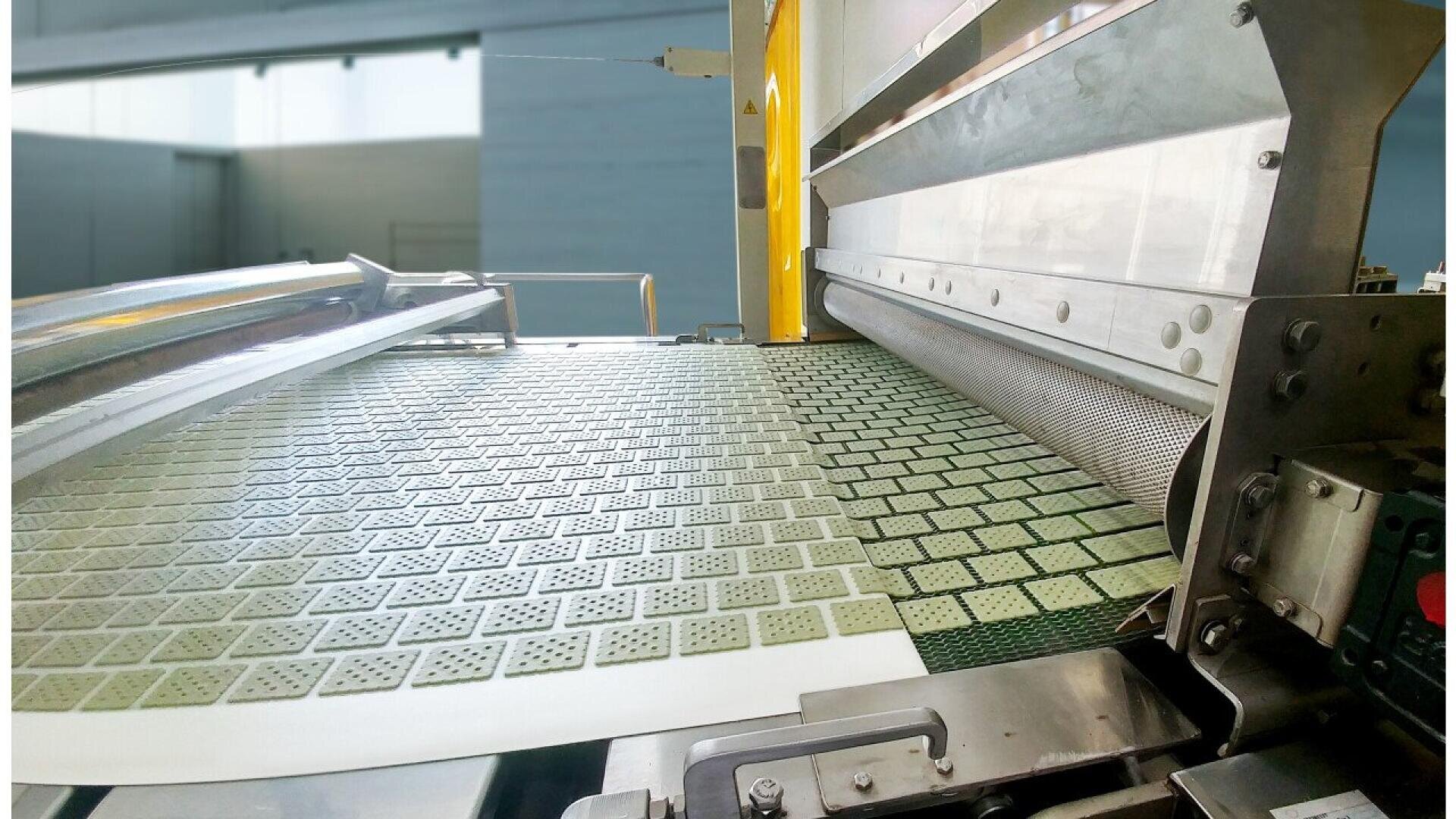 Nahaufnahme einer Industriemaschine, die Reihen von rechteckigen, perforierten Crackern oder Keksen auf einem Fließband in einer Fabrik produziert.
