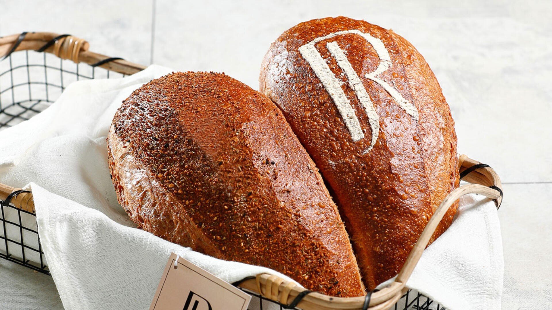 Zwei ovale Brotlaibe liegen in einem mit einem weißen Tuch ausgelegten Korb. Ein Laib hat eine strukturierte, genarbte Kruste, während der andere eine glatte Kruste mit einem weißen JR-Muster auf der Oberseite hat.