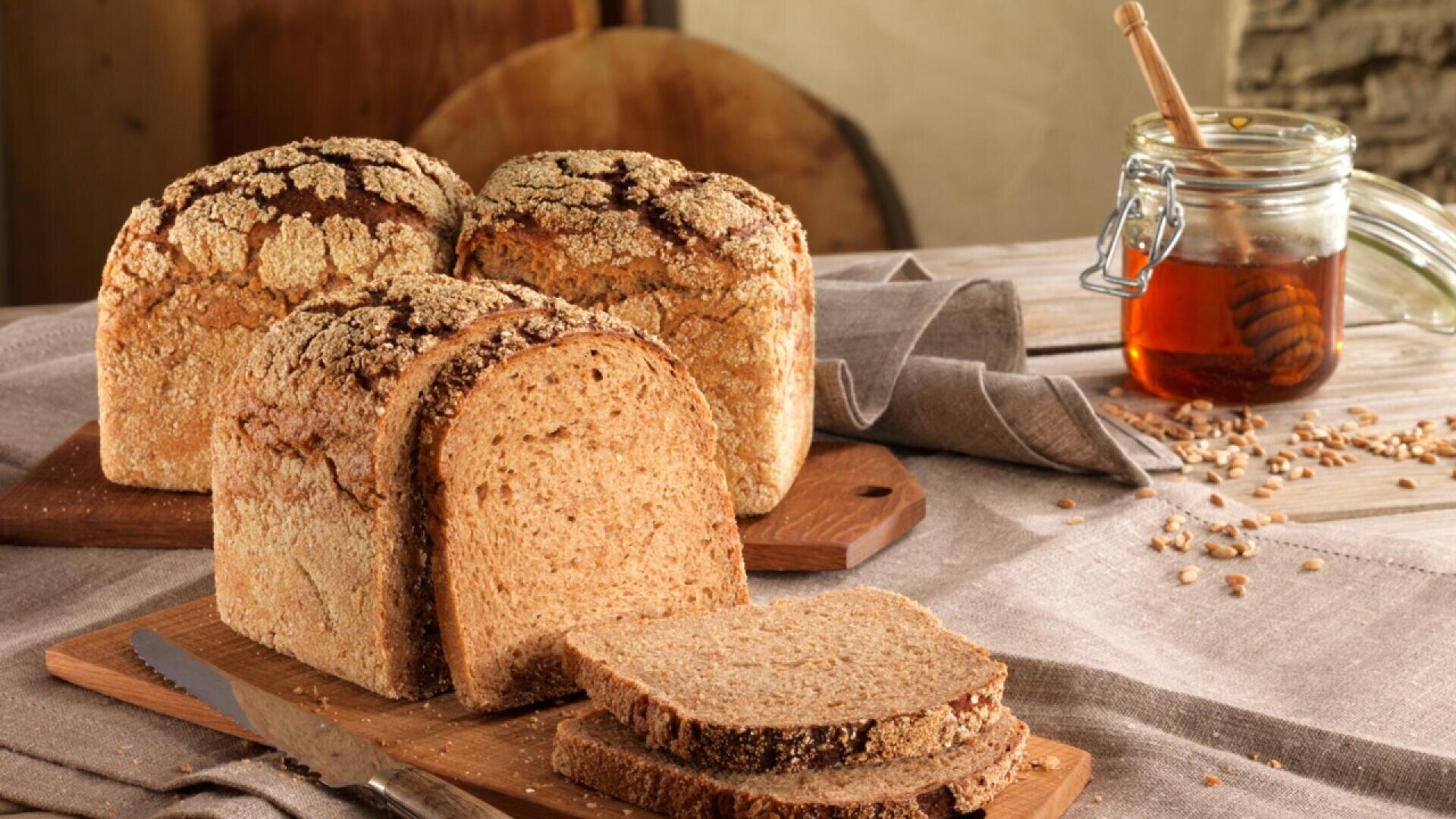 Three loaves of crusty whole grain bread sit on a wooden surface with several slices cut. A jar of honey with a dipper is nearby, along with scattered wheat grains and a knife.
