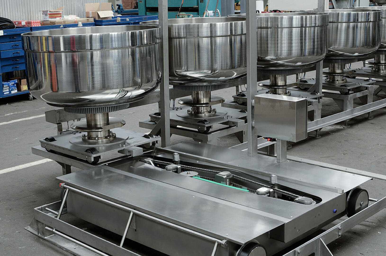 Row of large industrial stainless steel mixing bowls mounted on metal machinery in a factory setting, with tools and equipment visible in the background.