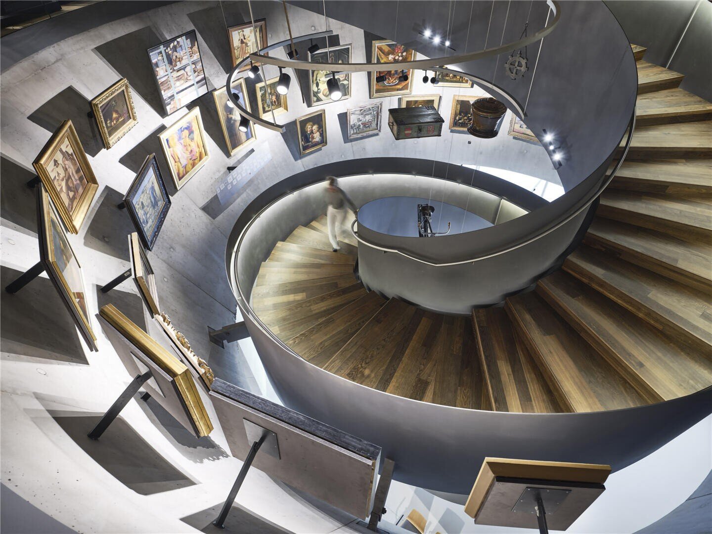 Eine Wendeltreppe mit hölzernen Stufen und einem Metallgeländer windet sich durch eine moderne Kunstgalerie mit gerahmten Gemälden an den geschwungenen Betonwänden und Staffeleien in der unteren Etage. Eine verschwommene Gestalt steigt die Treppe hinunter.