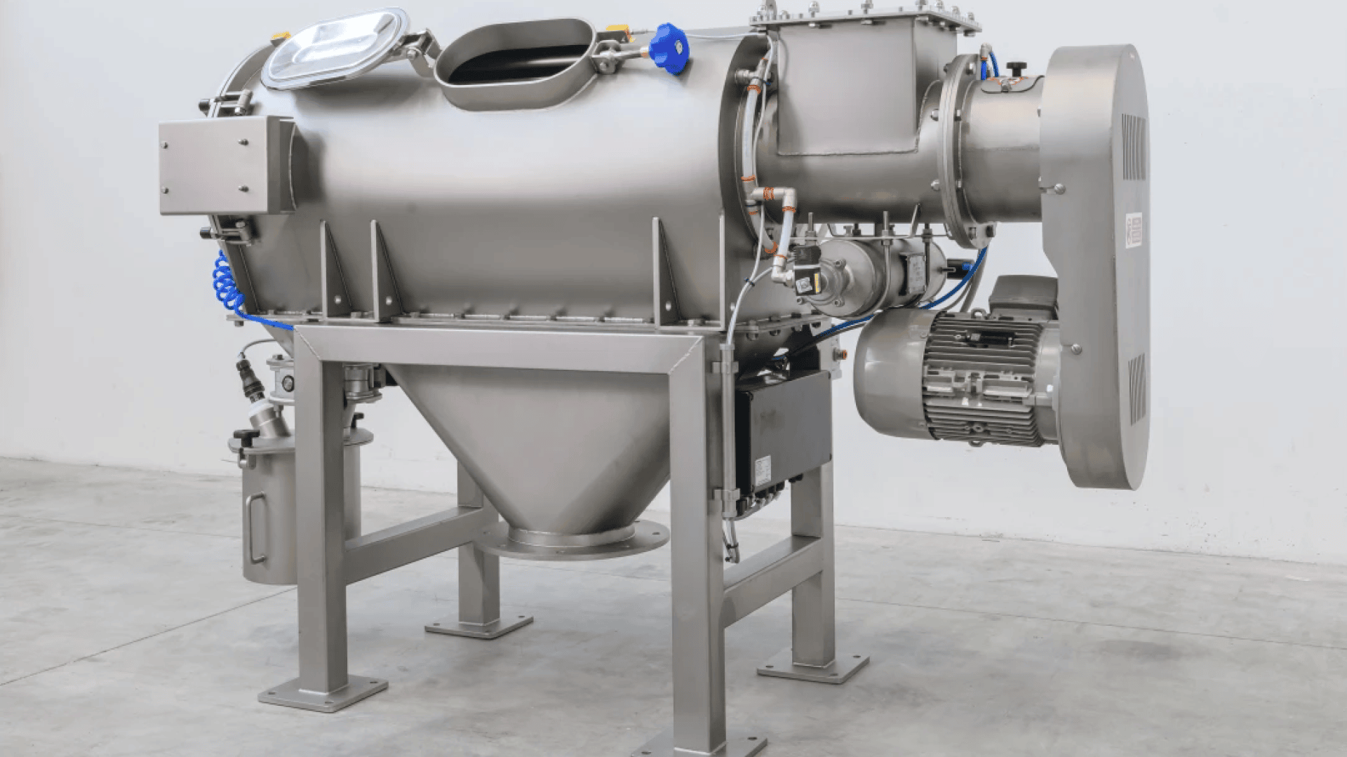 A large, industrial stainless steel machine with a hopper base, various pipes, valves, and a motor, positioned on a concrete floor against a plain white wall.