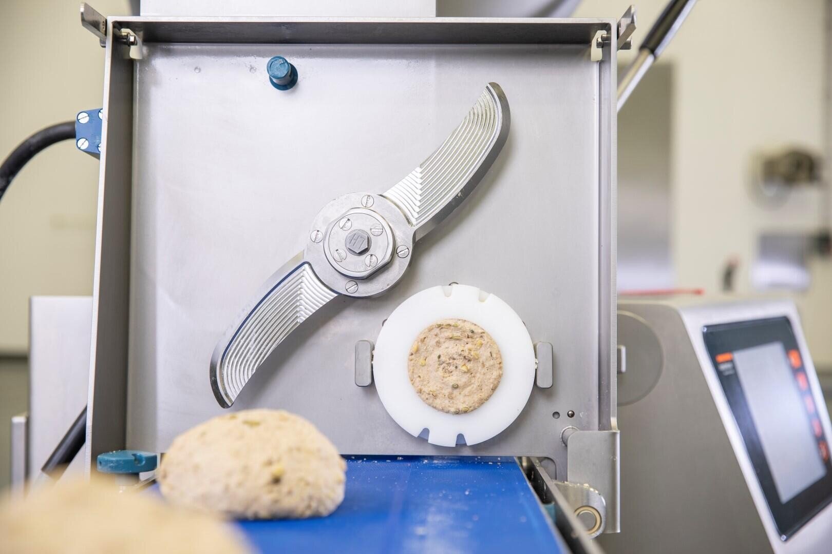 Nahaufnahme einer Lebensmittelverarbeitungsmaschine mit einer runden Ausstechform und einem plätzchenförmigen Teigstück auf einem weißen Tablett; ein weiteres Teigstück befindet sich auf einem blauen Förderband im Vordergrund.