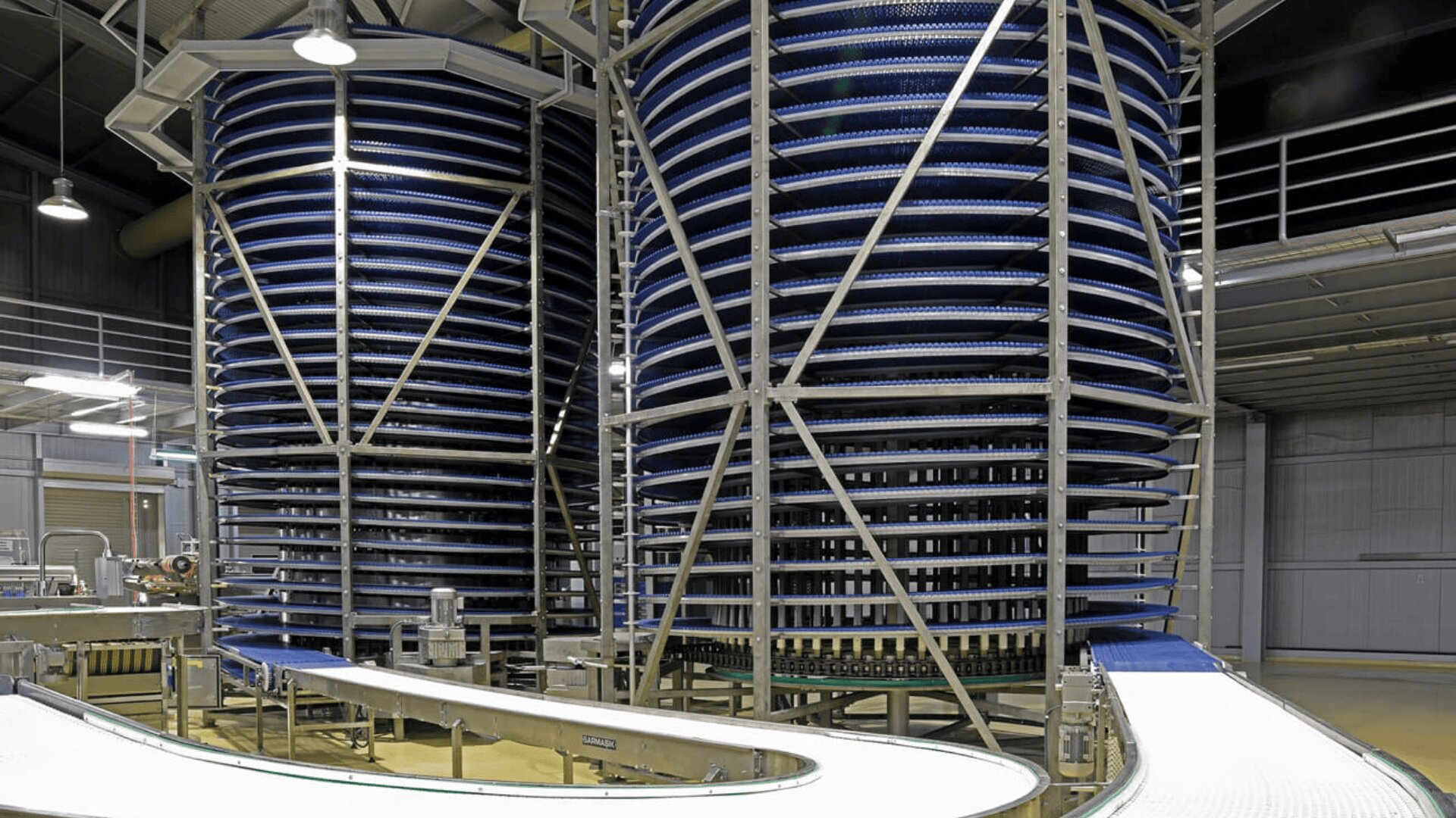 Large industrial spiral conveyor systems with blue belts are inside a modern factory, surrounded by metal beams and a white curved conveyor path in the foreground.