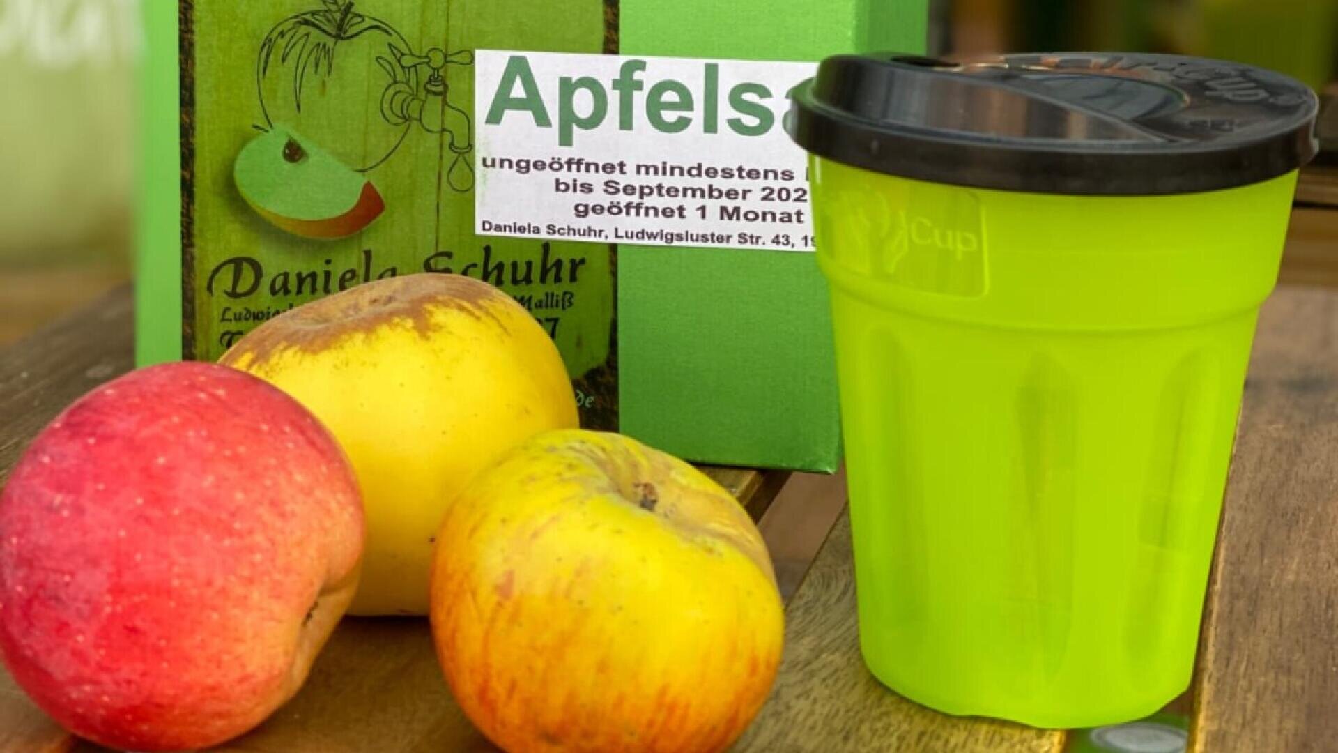 Three apples and a bright green reusable cup are on a wooden surface. Behind them is a green box labeled Apfels with additional German text and an illustration of an apple.