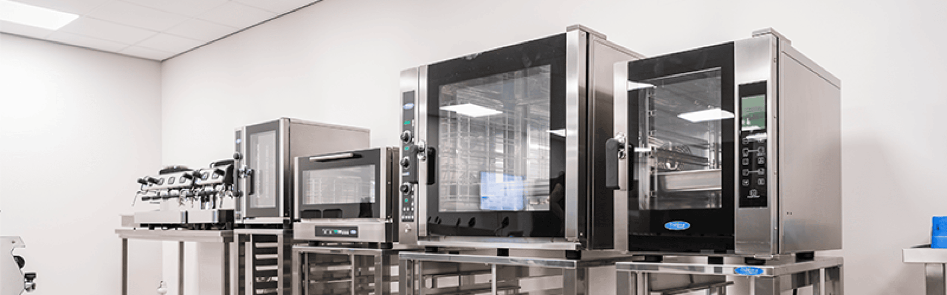 A row of commercial kitchen appliances, including two large stainless steel ovens, a microwave, and an espresso machine, set on metal counters in a clean, modern kitchen with white walls and ceiling.