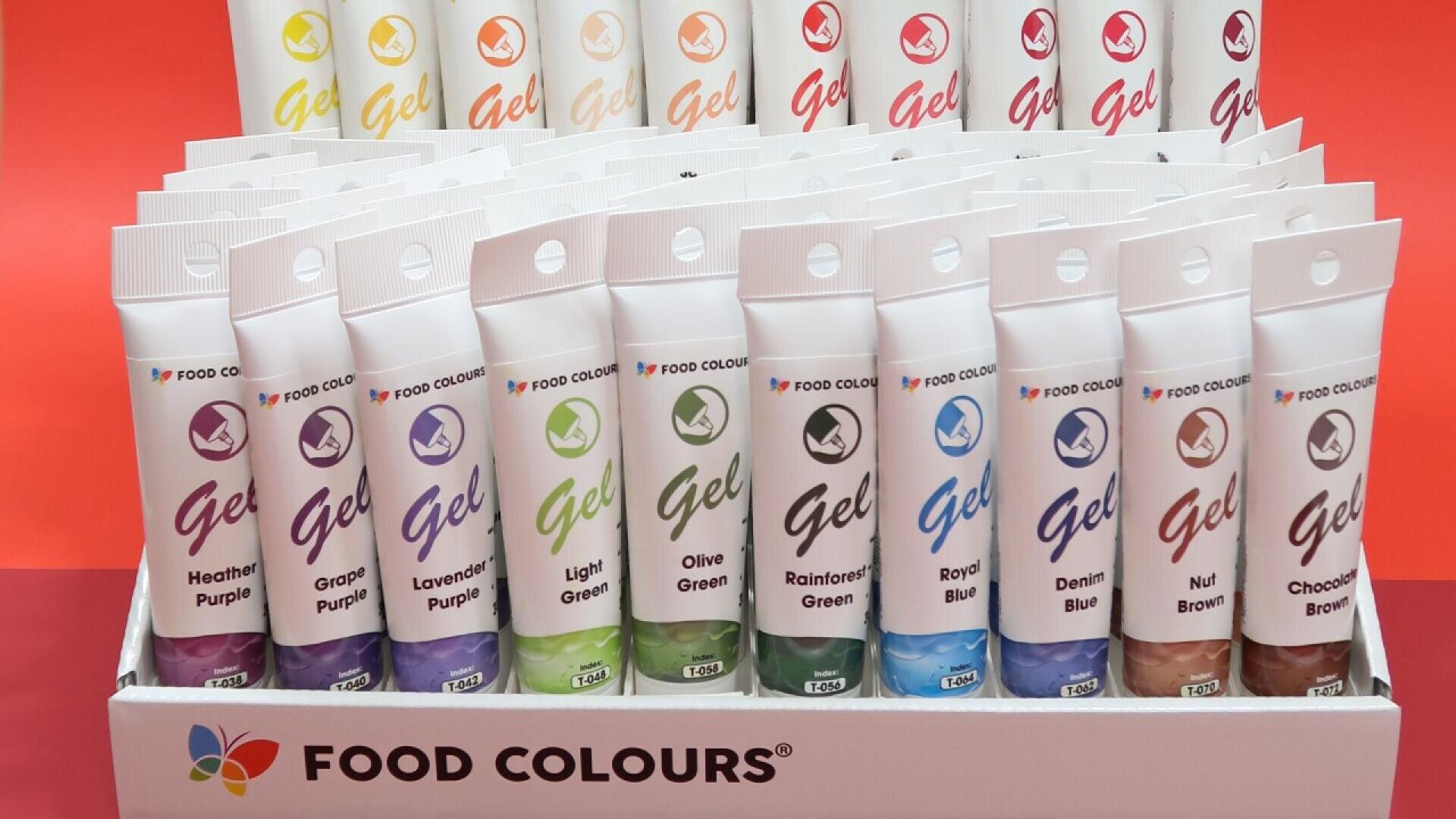 Ein Schaukasten mit Gel-Lebensmittelfarbtuben der Marke FOOD COLOURS in verschiedenen Farben, darunter lila, grün, blau und braun, die in Reihen vor einem roten Hintergrund angeordnet sind.