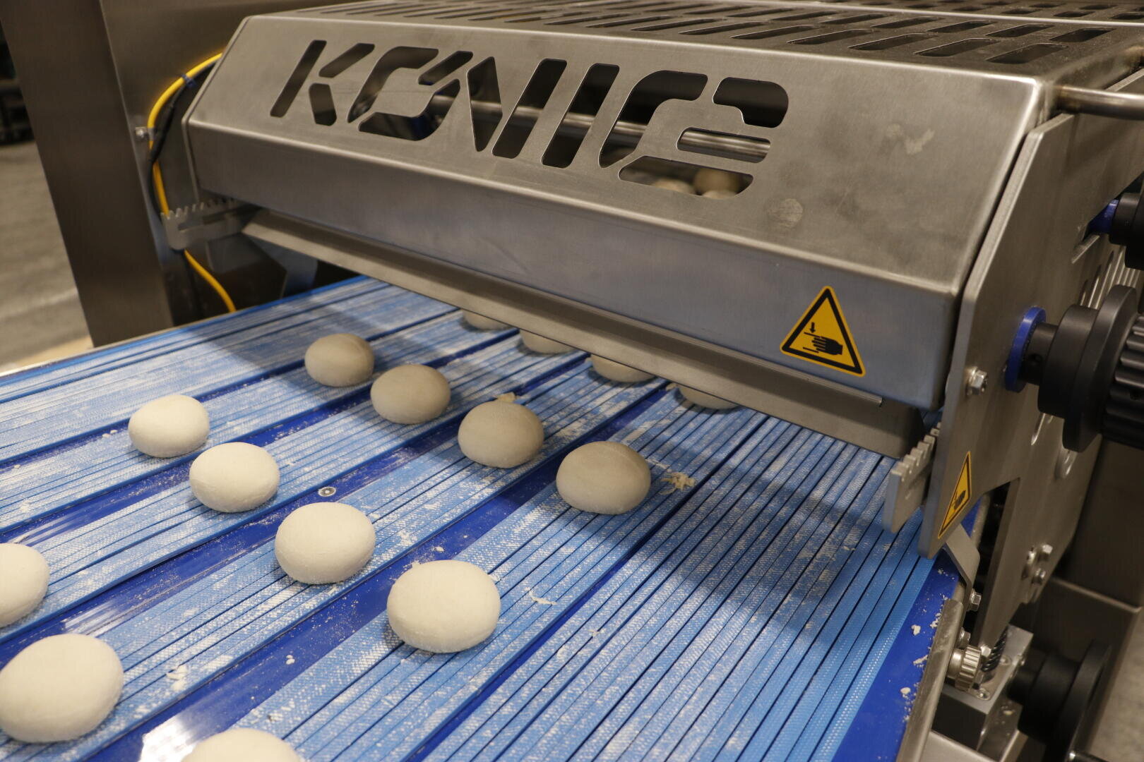 A metal machine labeled KONIG shapes dough balls on a blue conveyor belt, with a caution sign visible on the side. The dough balls are evenly spaced and moving along the belt.