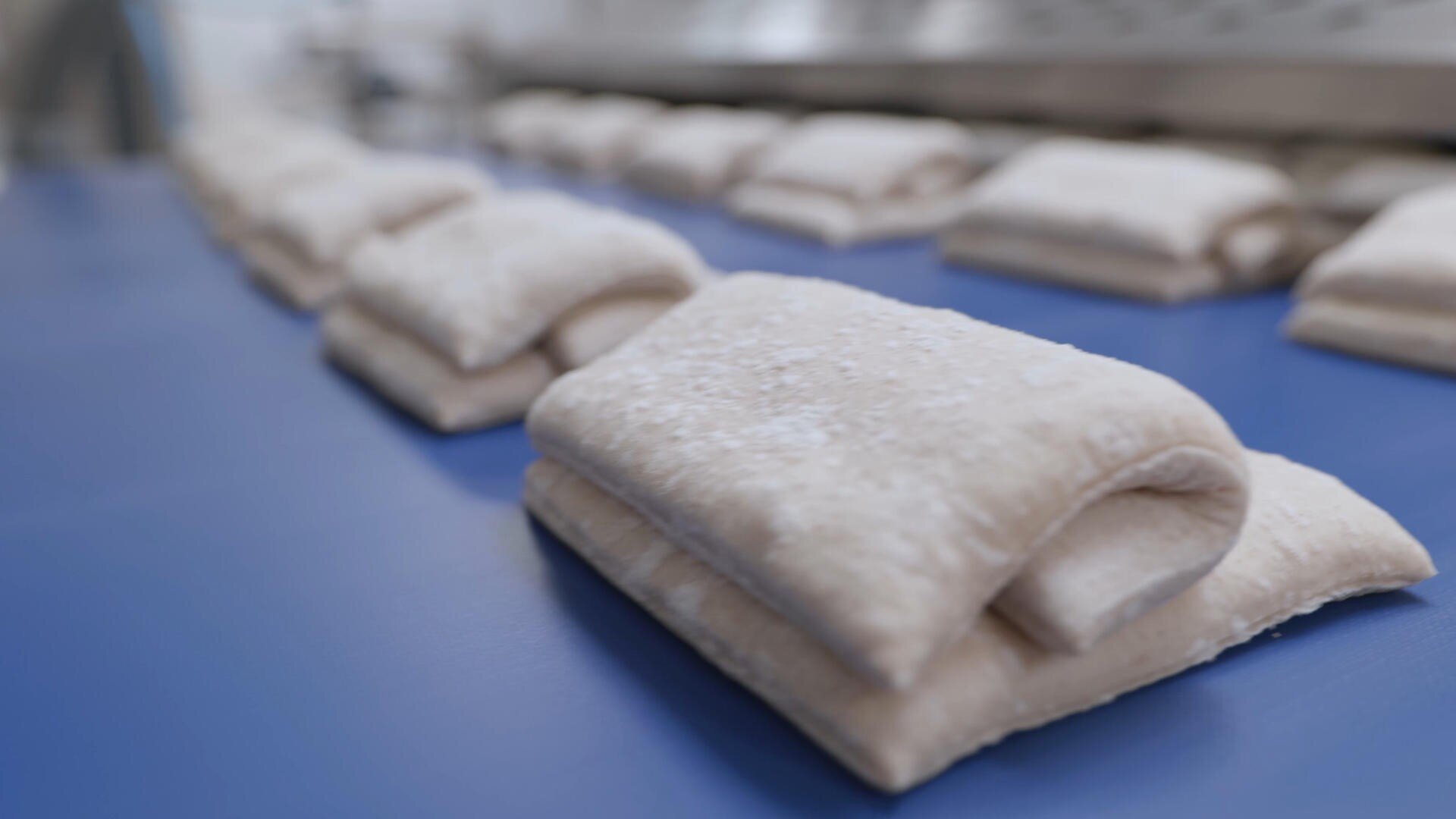 Auf einem blauen Förderband werden in einer Fabrikanlage Stapel von gefalteten Teiglingen aufgereiht, die zur Weiterverarbeitung oder zum Backen bereit sind.