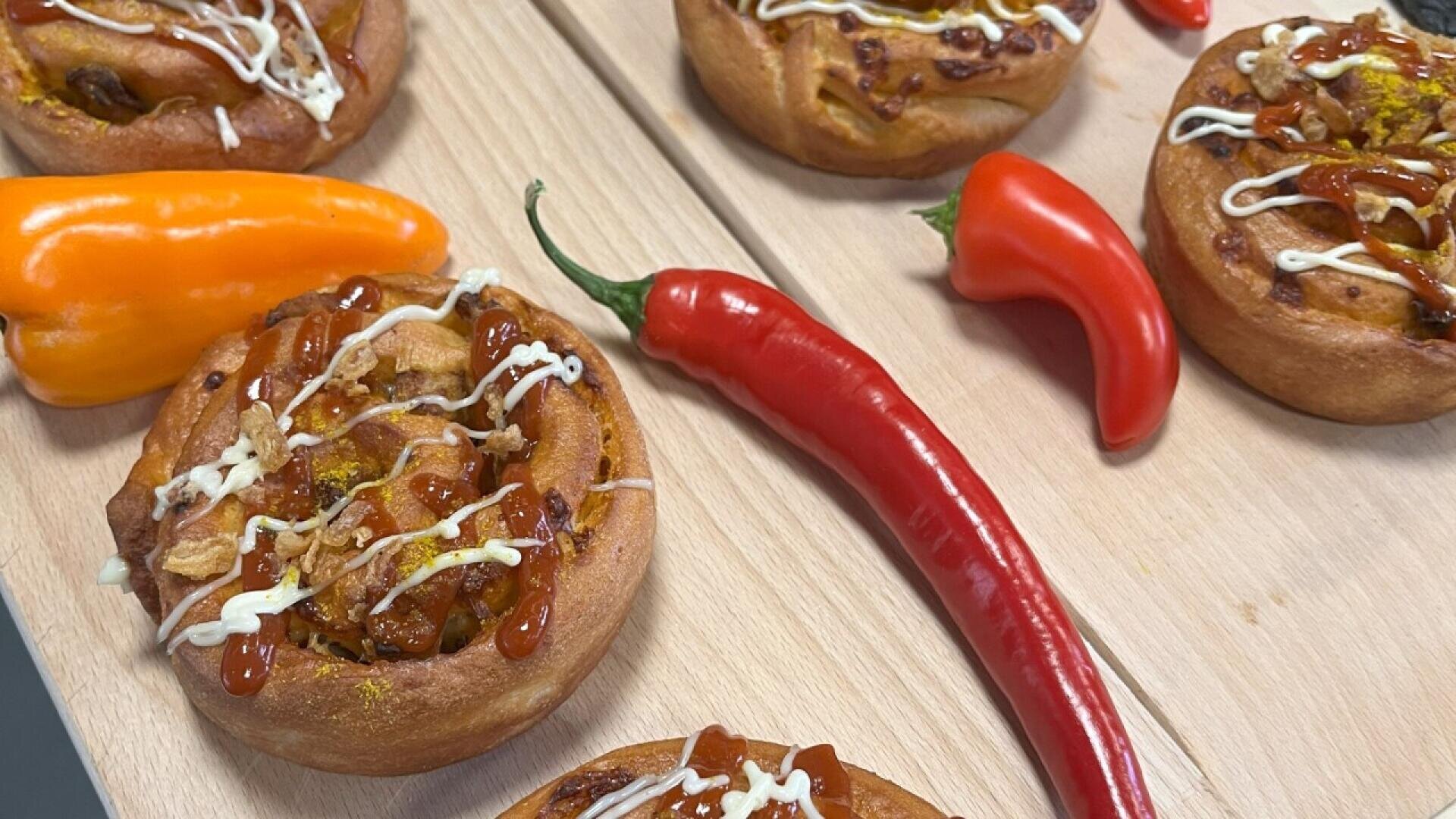 Savory baked rolls topped with sauce and drizzle, arranged on a wooden surface alongside whole orange, red, and long red chili peppers.