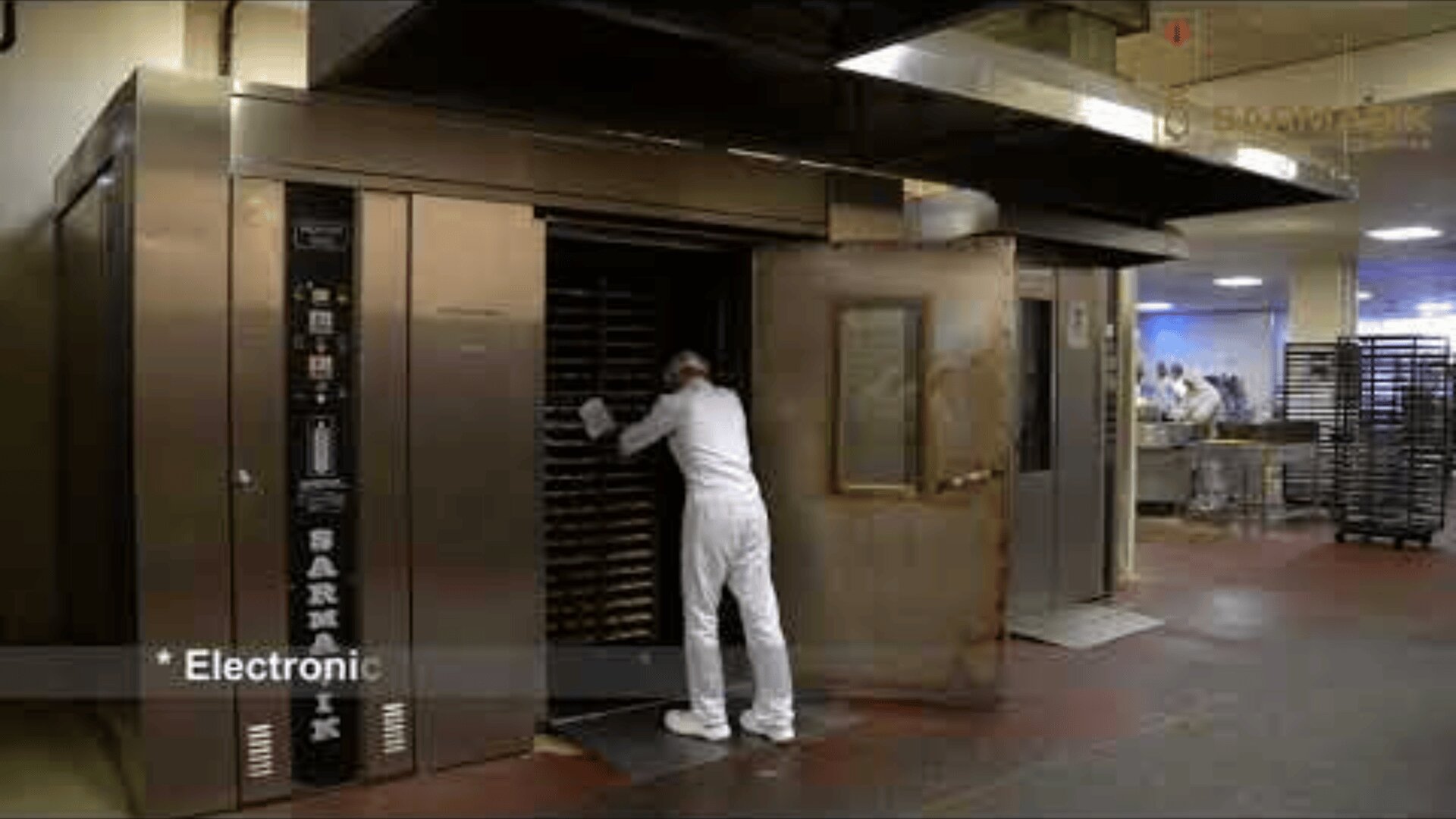 A worker in white uniform loads a large industrial oven with trays in a commercial bakery kitchen; another group works in the background. The word Electronic appears in the lower left corner.