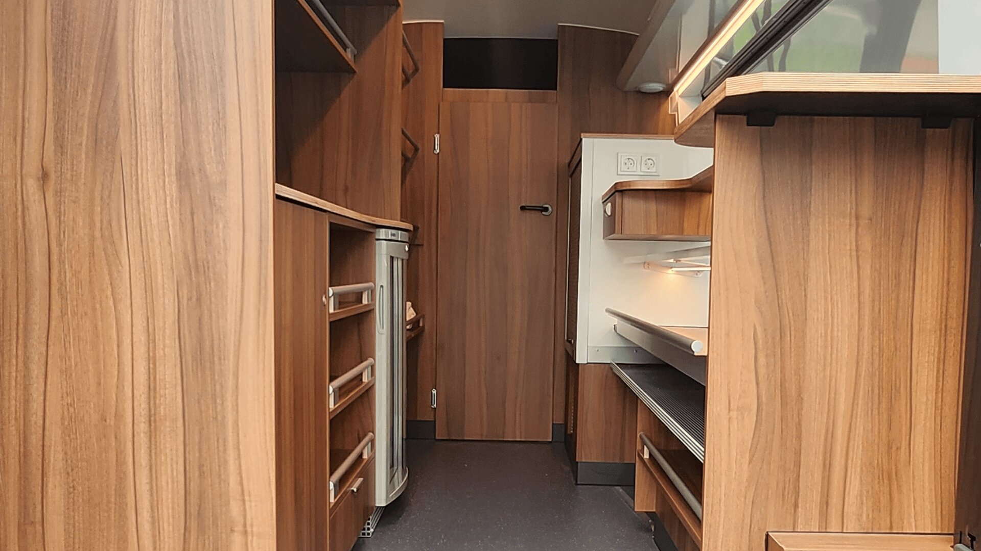 Innenraum eines kompakten Raums oder Wohnmobils mit Holzschränken, Schubladen und Regalen auf beiden Seiten, einer Arbeitsplatte mit einem Ofen und einer geschlossenen Holztür am anderen Ende. Der Bodenbelag ist dunkel und die Beleuchtung ist gedämpft.