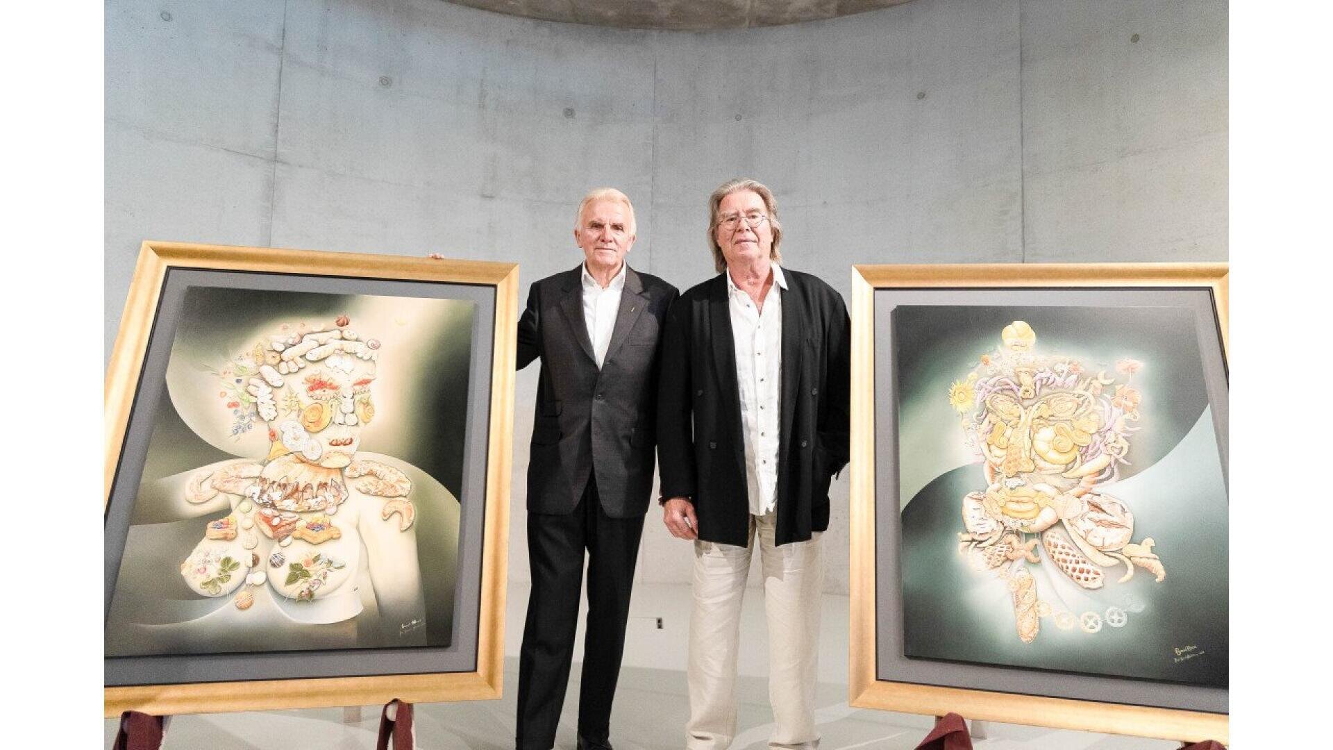 Two men in suits stand side by side indoors, each with a hand on a large, framed abstract artwork featuring intricate, colorful designs. The artworks are displayed on easels in a modern, minimalist room.