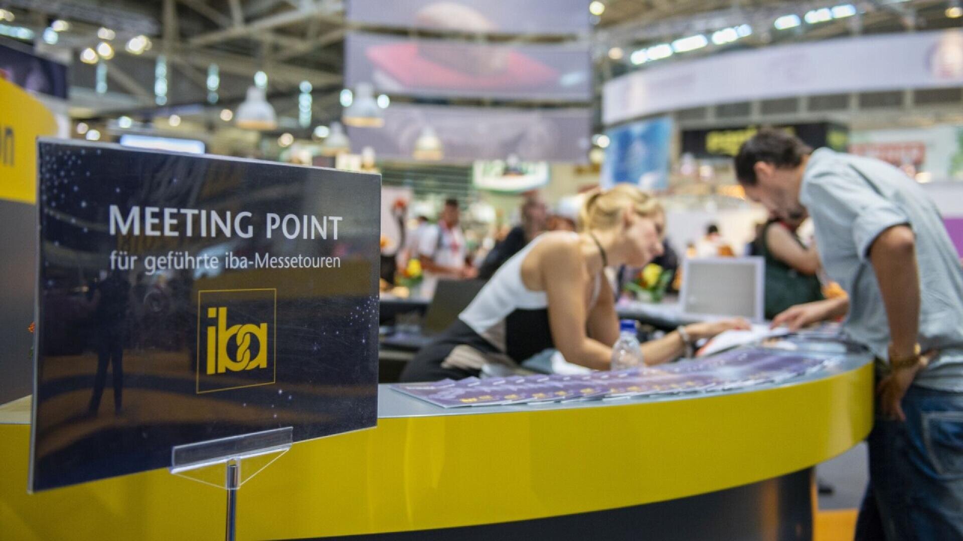 People gather at a curved information counter in a busy exhibition hall. A sign in the foreground reads “MEETING POINT für geführte iba-Messetouren” with the iba logo. The background shows various booths and displays.