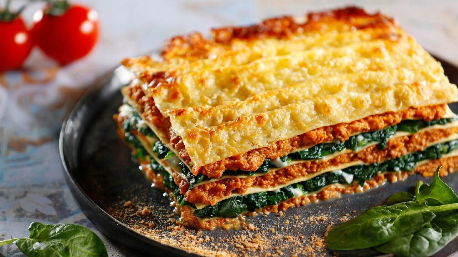 Eine Nahaufnahme eines Stücks Lasagne mit Schichten aus Nudeln, Spinat, Käse und Tomatensoße auf einem schwarzen Teller, garniert mit frischen Spinatblättern und Tomaten im Hintergrund.