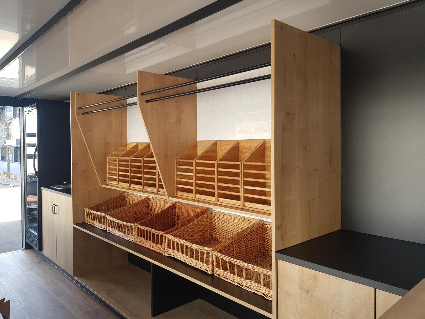 Holzregale und Fächer mit mehreren leeren Weidenkörben sind in einem modernen, gut beleuchteten Laden oder Marktstand mit schwarzen Arbeitsplatten und weißen Kacheln übersichtlich angeordnet.