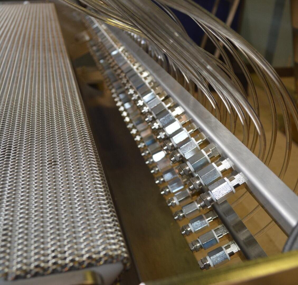 Nahaufnahme eines metallischen Industriemaschinenteils mit einer Netzoberfläche auf der linken Seite und Reihen verbundener Metallbeschläge und transparenter Rohre auf der rechten Seite.