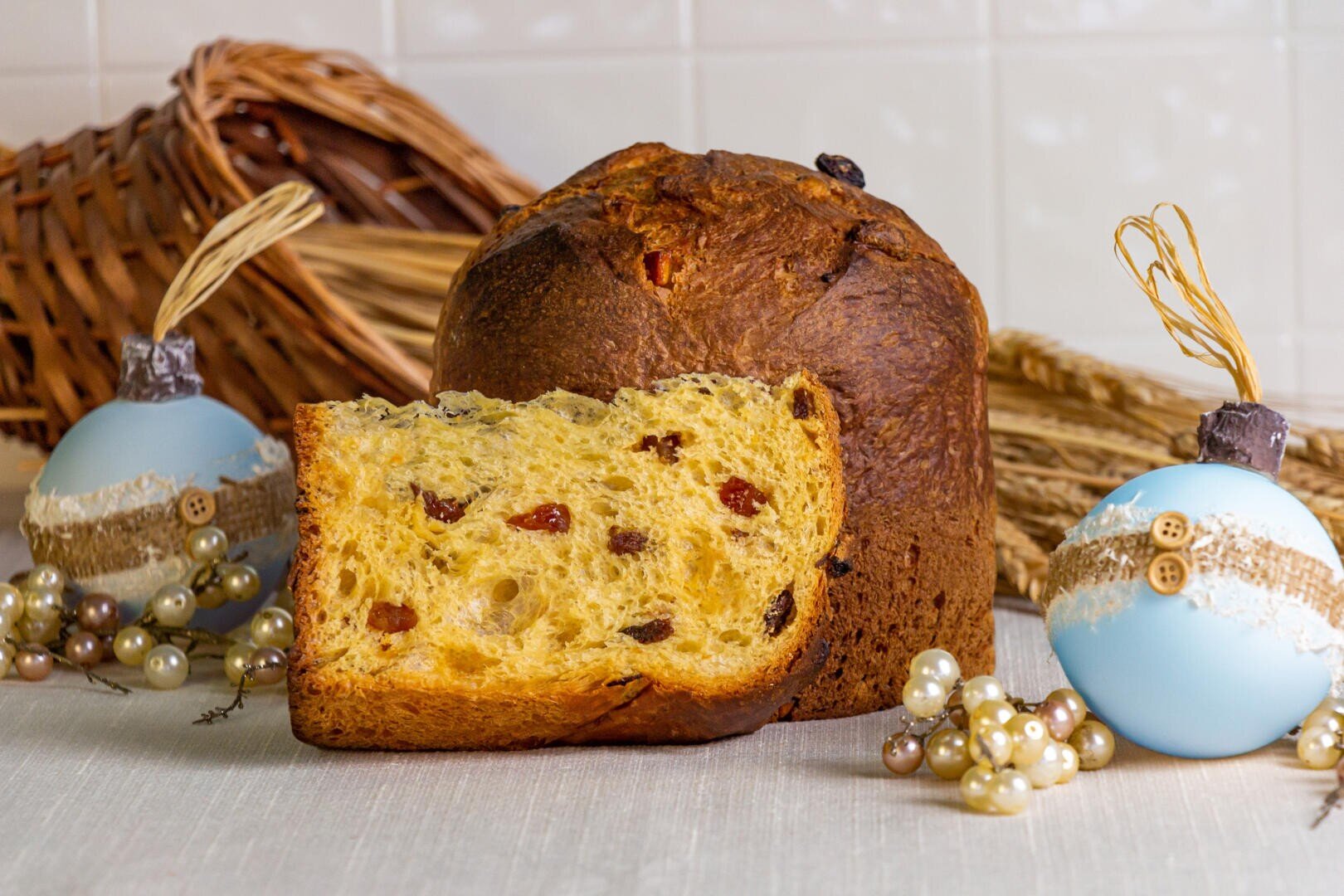 A sliced loaf of panettone with visible raisins sits on a table, surrounded by light blue Christmas ornaments, pearl garlands, and a wicker basket in the background.