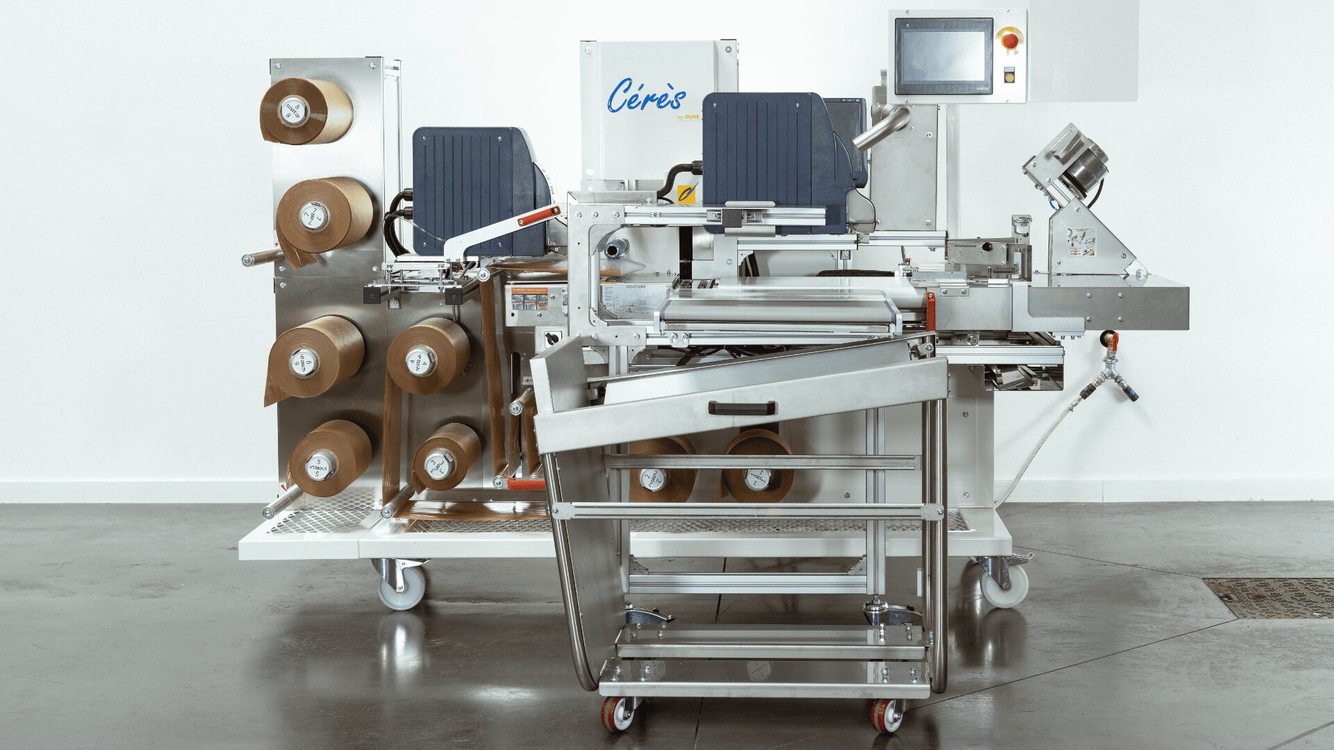 A stainless steel industrial packaging machine with multiple brown paper rolls, control panels, and touch screen, set on wheels in a clean, white room.