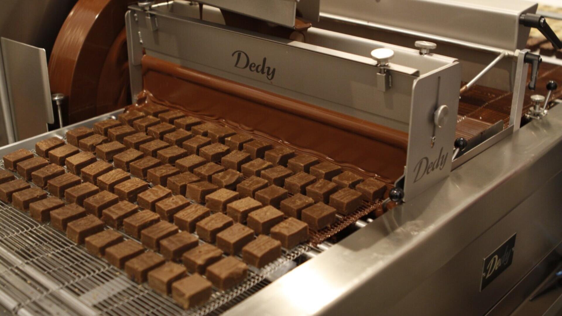 A machine labeled “Dedy” coats rows of square chocolates with liquid chocolate, moving them along a conveyor belt in a factory setting.