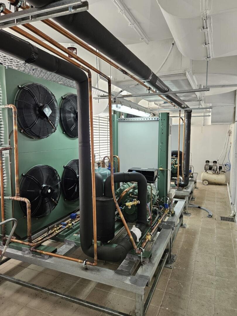 Two large industrial refrigeration units with visible copper pipes, black insulation, and big cooling fans are installed in a clean, well-lit mechanical room. An air compressor is seen in the background.