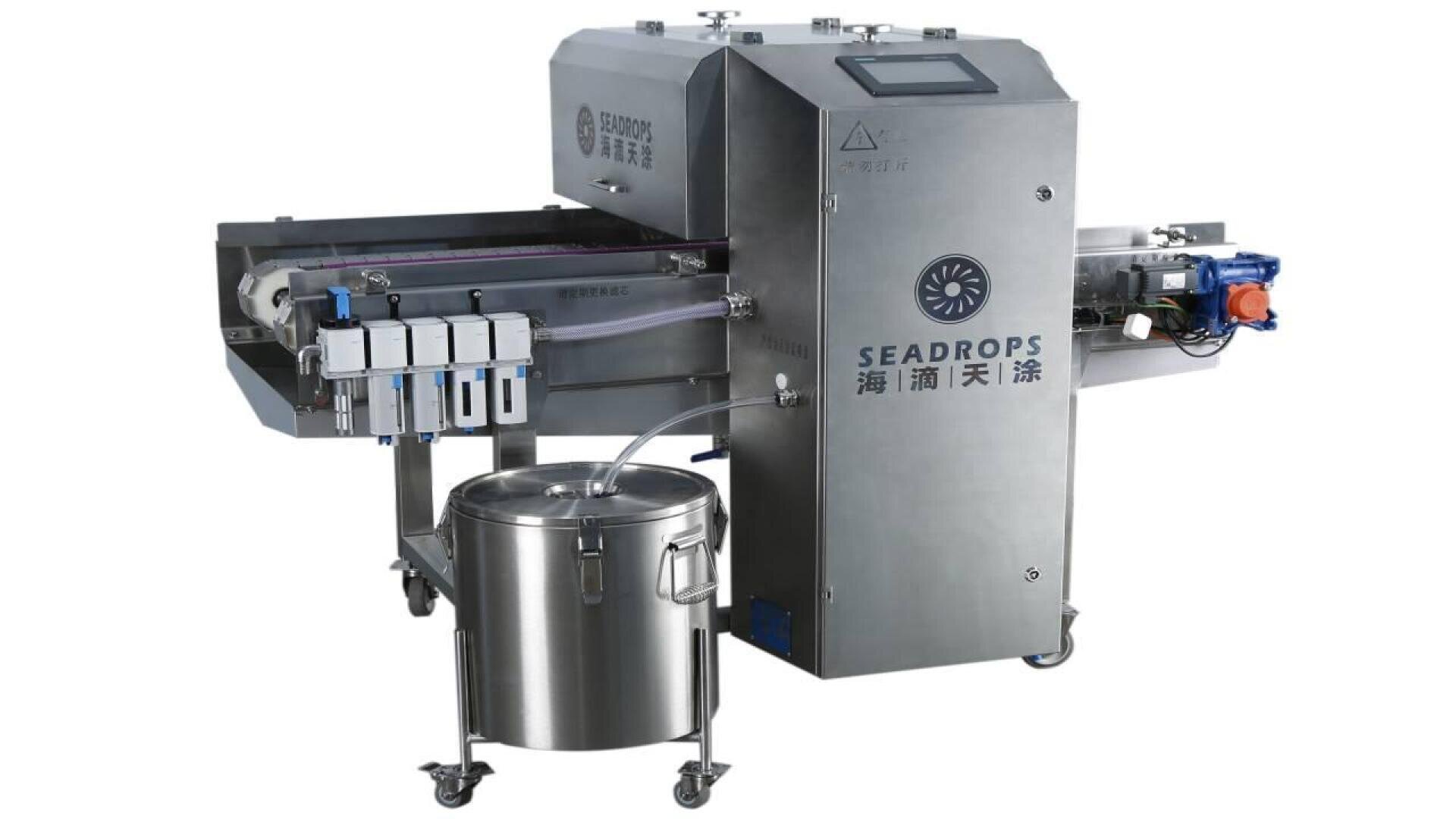 A stainless steel industrial food processing machine labeled Seadrops, featuring a conveyor belt, digital control panel, four dispensers, and a large cylindrical container on wheels beneath the output area.